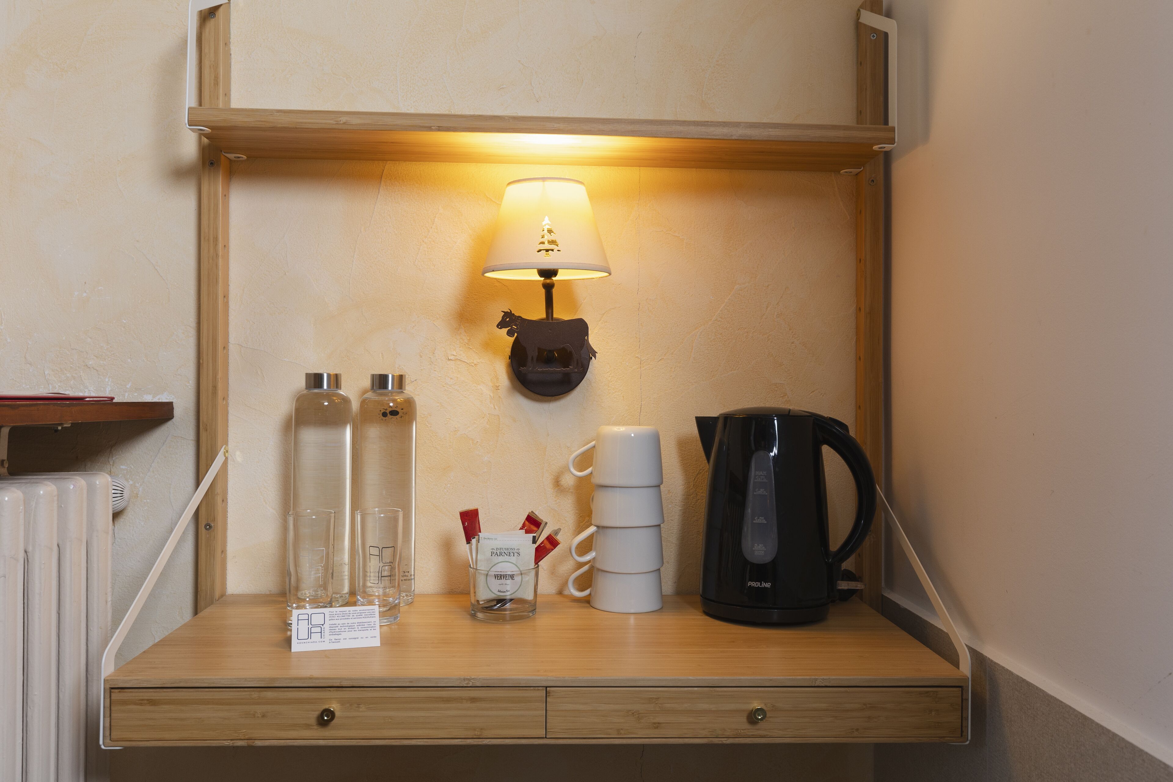 A wooden shelf with a lamp, a stack of white cups, a black coffee maker, and two glass bottles with a small card.