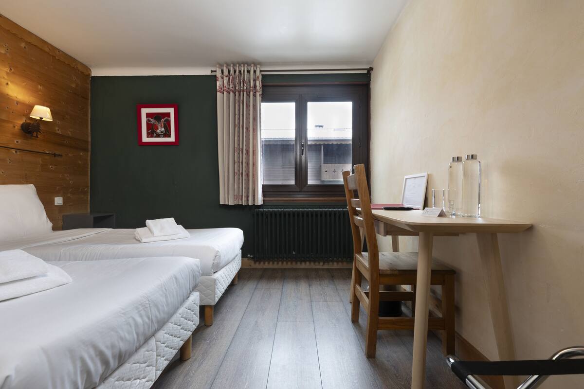 Ein Hotelzimmer mit zwei Betten, einem Holztisch, einem Stuhl, einem Heizkörper und einem Fenster mit Vorhängen.