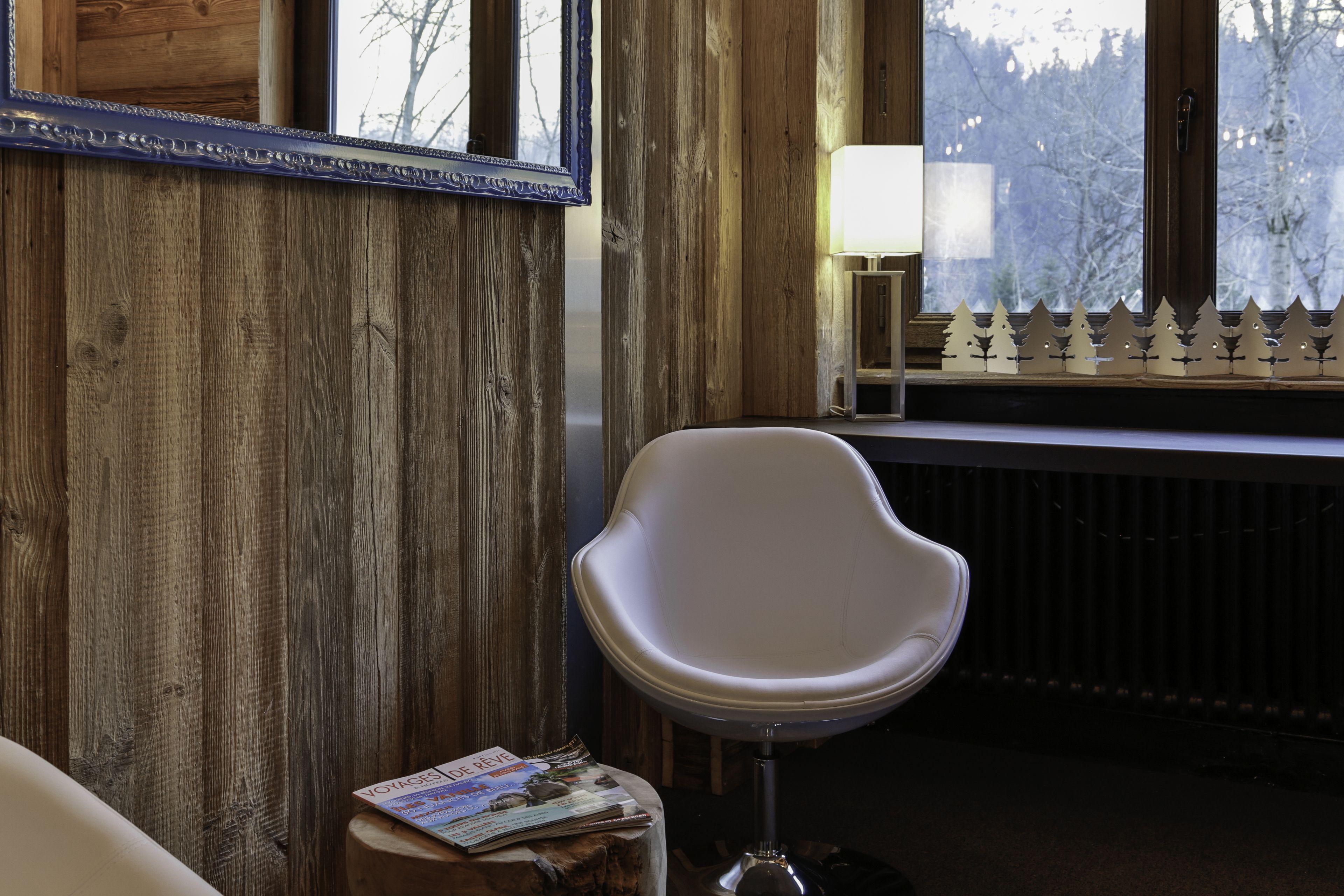 Una habitación con paredes de madera, una silla blanca, una revista sobre un tocón de madera y una ventana con vistas a árboles.