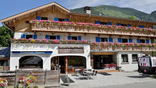 Ein Hotel mit Restaurant und Pizzeria im Erdgeschoss, mit Balkonen, bepflanzten Blumenkästen und Bergblick.