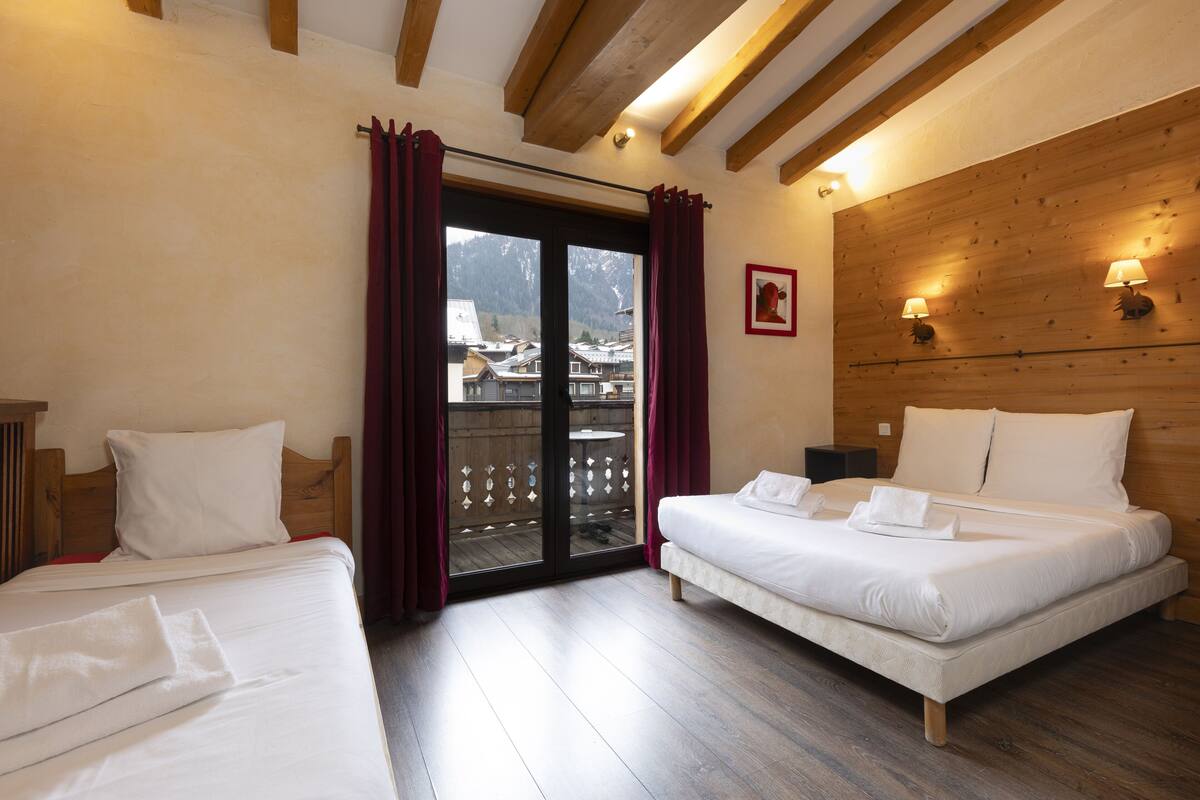 Ein Hotelzimmer mit zwei Betten, Holzbalkendecke, einem Balkon mit Blick auf ein Bergdorf und Wandlampen.