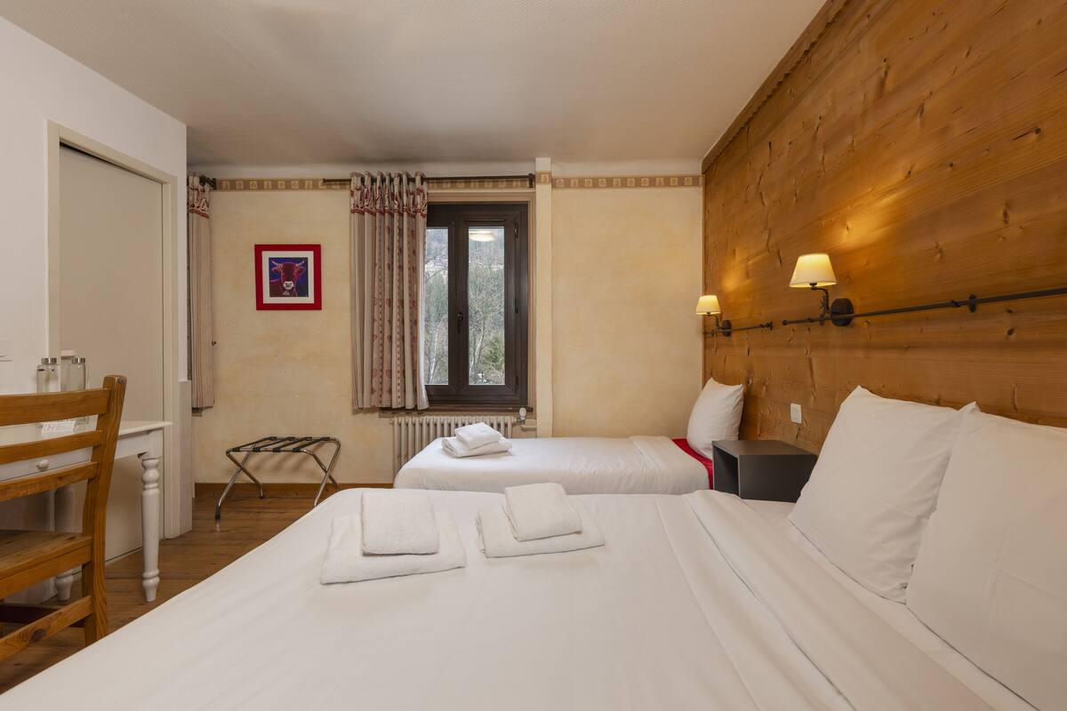 Ein Hotelzimmer mit zwei Betten, einem Holzkopfstück, einem Fenster mit Vorhängen, einem Schreibtisch mit Stuhl und einem Heizkörper.