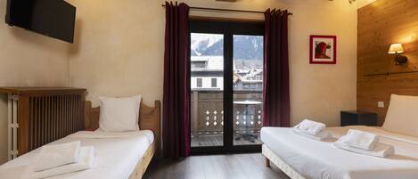 Habitación de hotel con dos camas, un televisor montado en la pared, techo de madera y vistas a edificios a través de un ventanal.