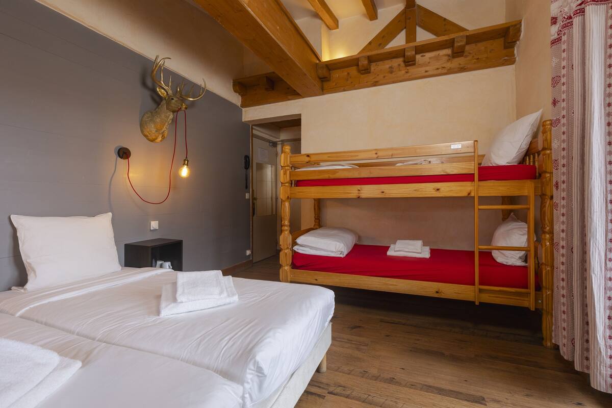 Ein Zimmer mit zwei Betten, eines mit roter und eines mit weißer Matratze. Am Kopfteil des Bettes befindet sich ein hölzerner Hirschkopf. Ein kleiner Nachttisch mit Lampe ist zu sehen.