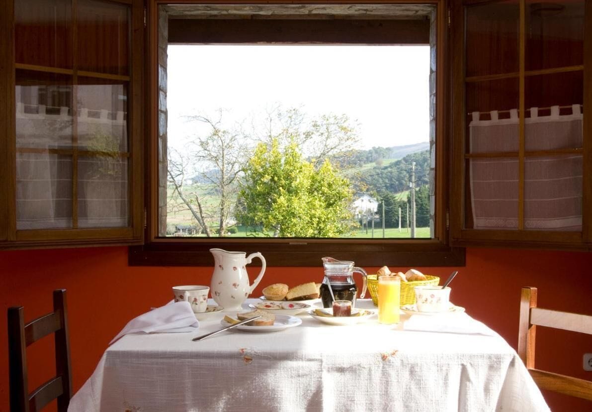 Mesa de café da manhã com jarra, xícaras e cesta de pão, perto de uma janela com vista para árvores e colinas.