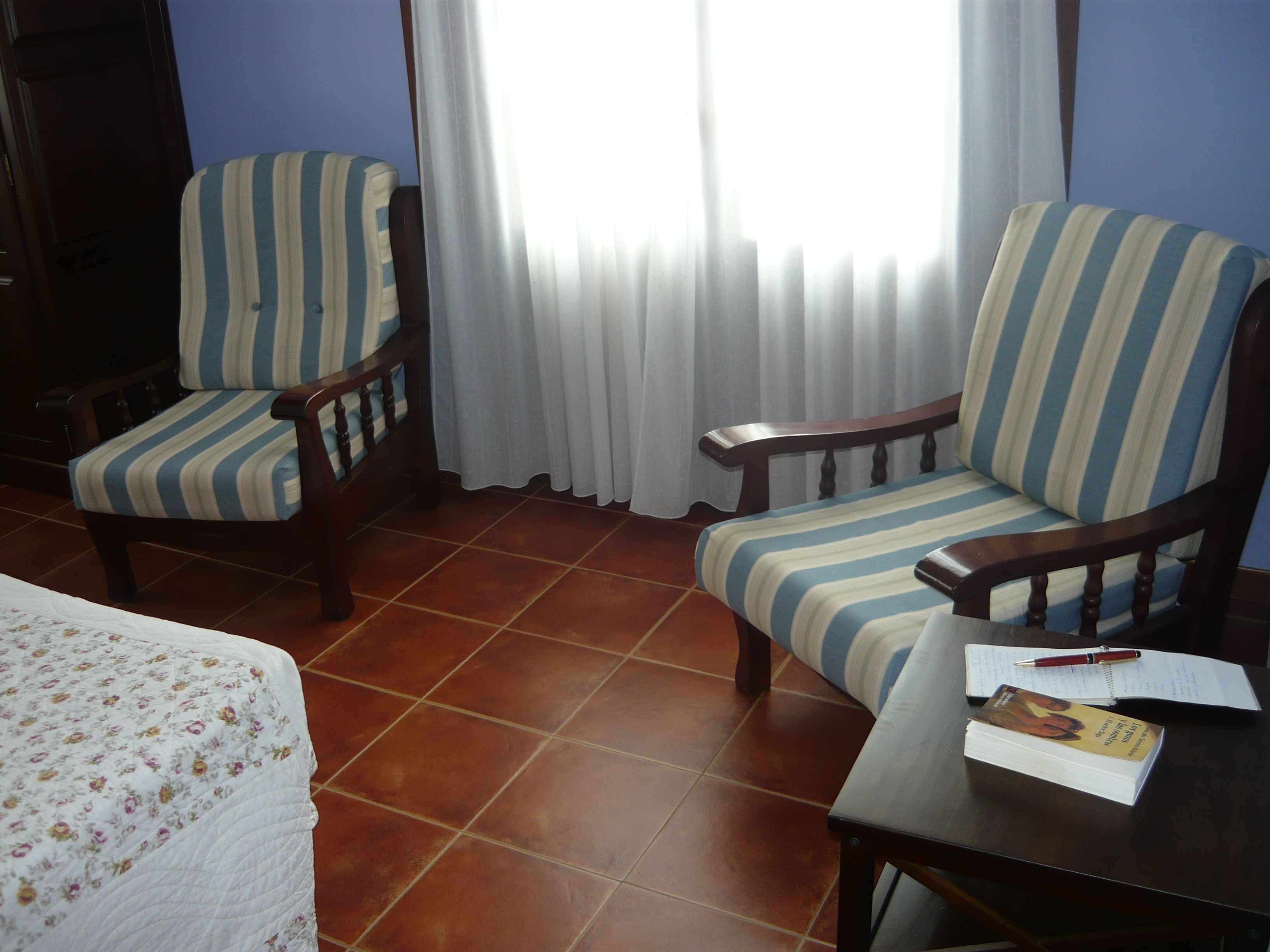 Una habitación con dos sillones a rayas, una mesa de madera con un libro y un bolígrafo, y una cama con colcha estampada.