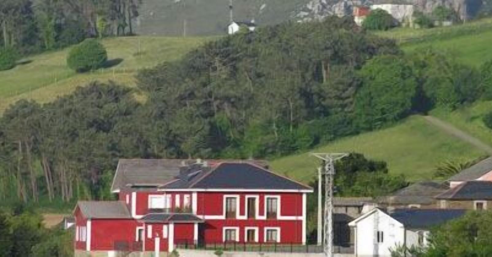 A red house with a dark roof, surrounded by greenery and hills.