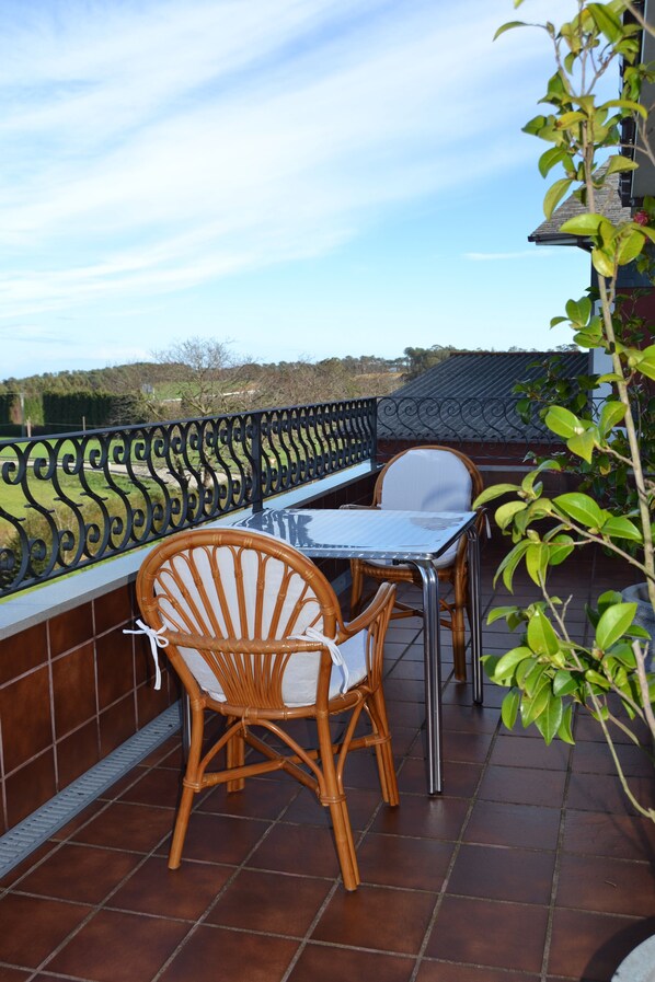 A balcony with a table and two wicker chairs, a metal railing, and a view of a green landscape.
