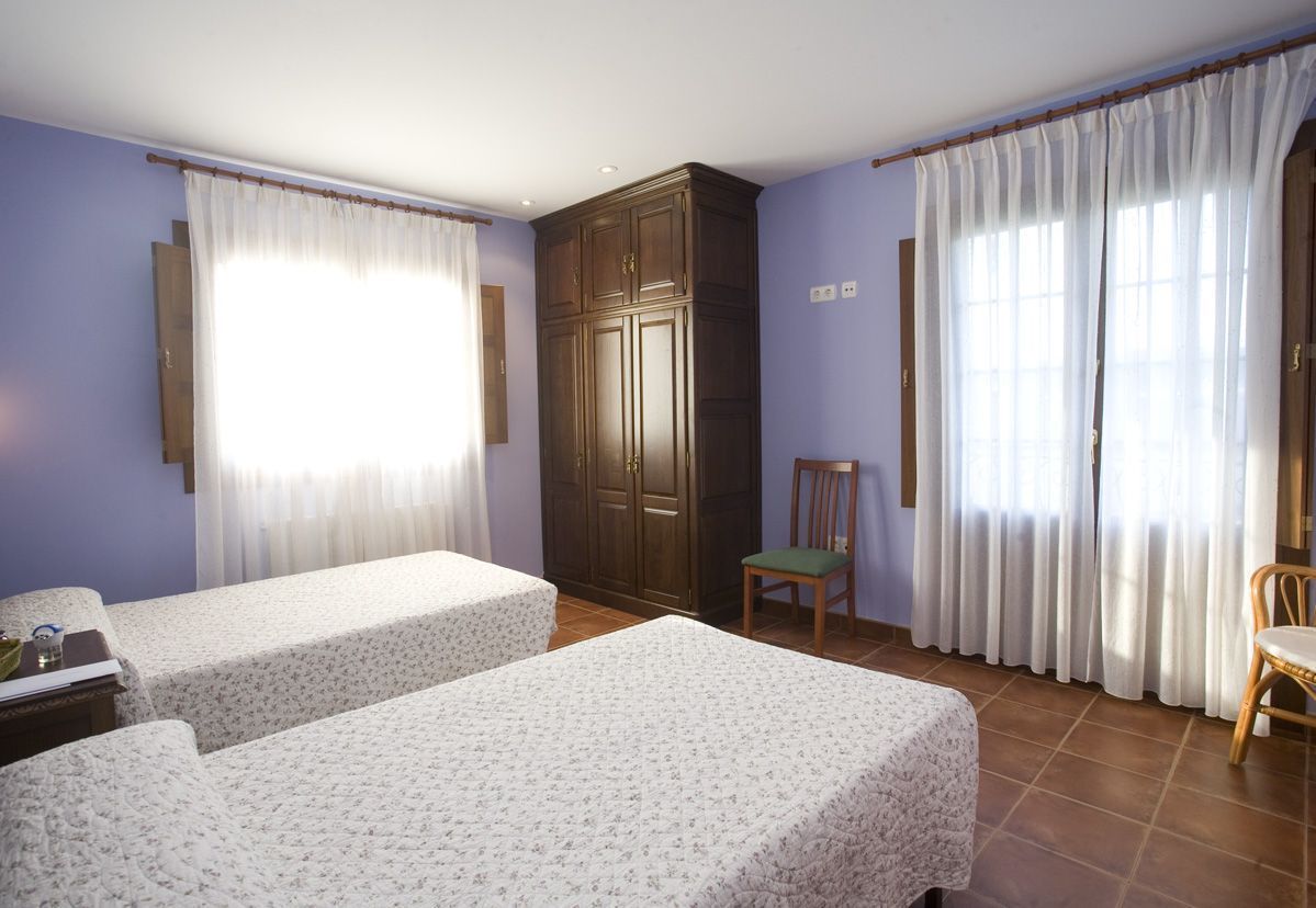 Una habitación con dos camas, un armario, una silla y ventanas amplias con cortinas.