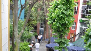 Um beco estreito de paraleipípedos, com mesas ao ar livre sob guarda-sóis, cercado por vegetação e edifícios.