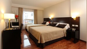Quarto de hotel com duas camas, uma escrivaninha com um laptop, uma luminária e vista da cidade pela janela.