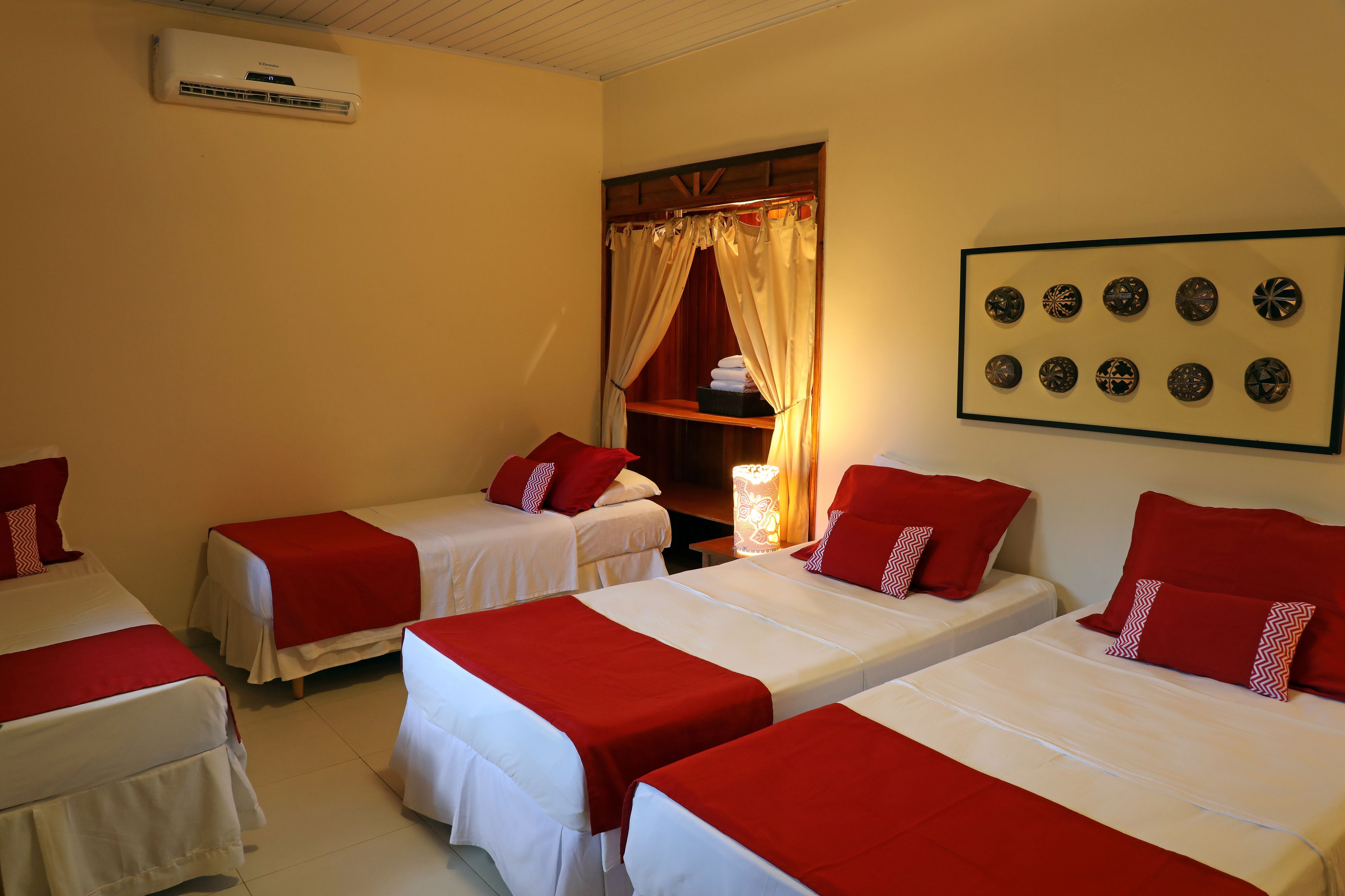 A hotel room with three single beds, each with red and white bedding, a wall-mounted air conditioner, and a framed collection of decorative plates on the wall.