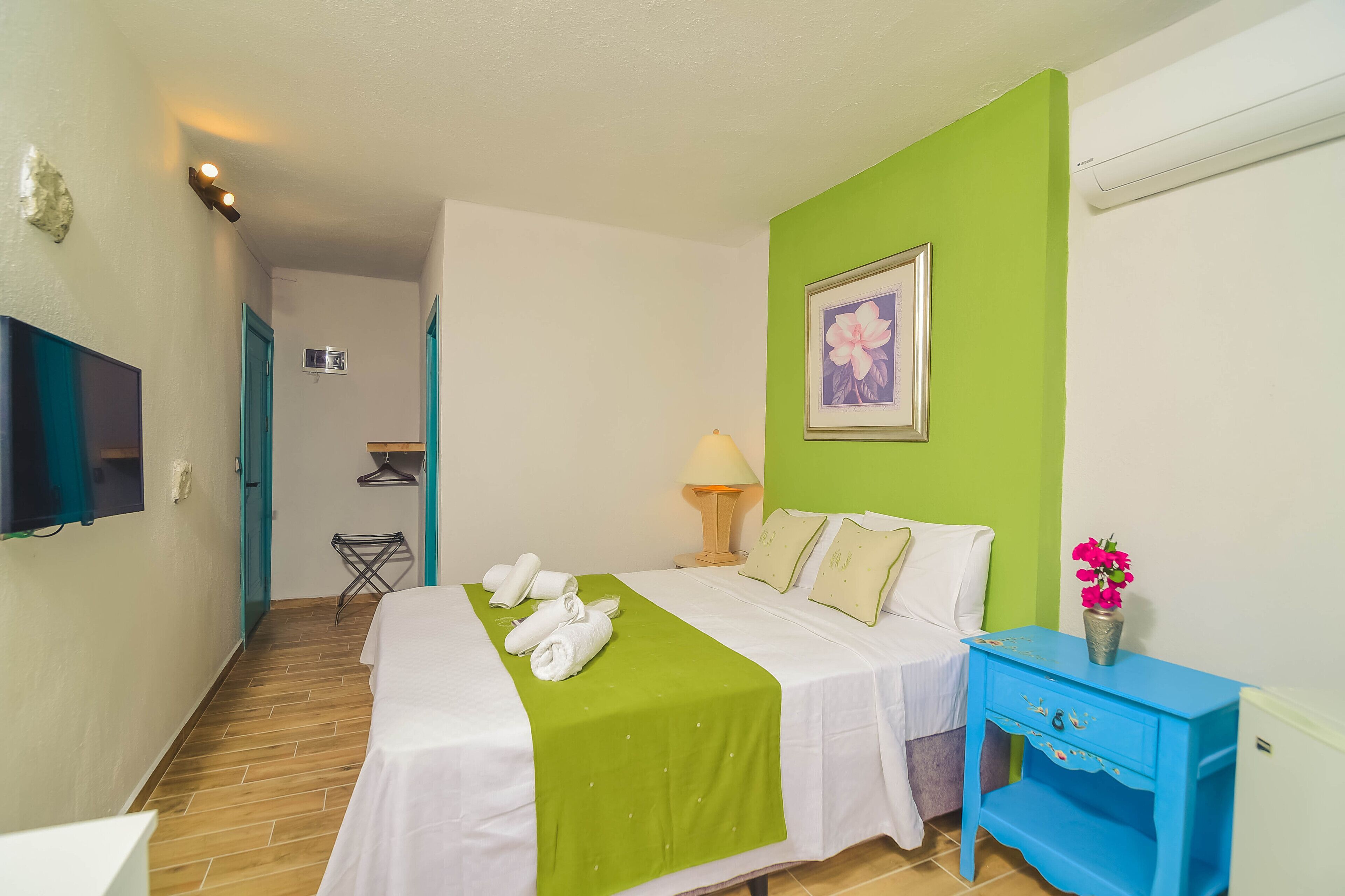 Een hotelkamer met een bed, een blauwe nachtkastje, een tv aan de muur, een groene accentmuur en een ingelijst beeld.