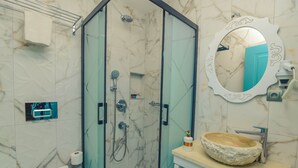 Un baño moderno con ducha envuelta en vidrio, pared con mosaicos de mármol, espejo redondo con marco decorativo y lavabo de piedra.