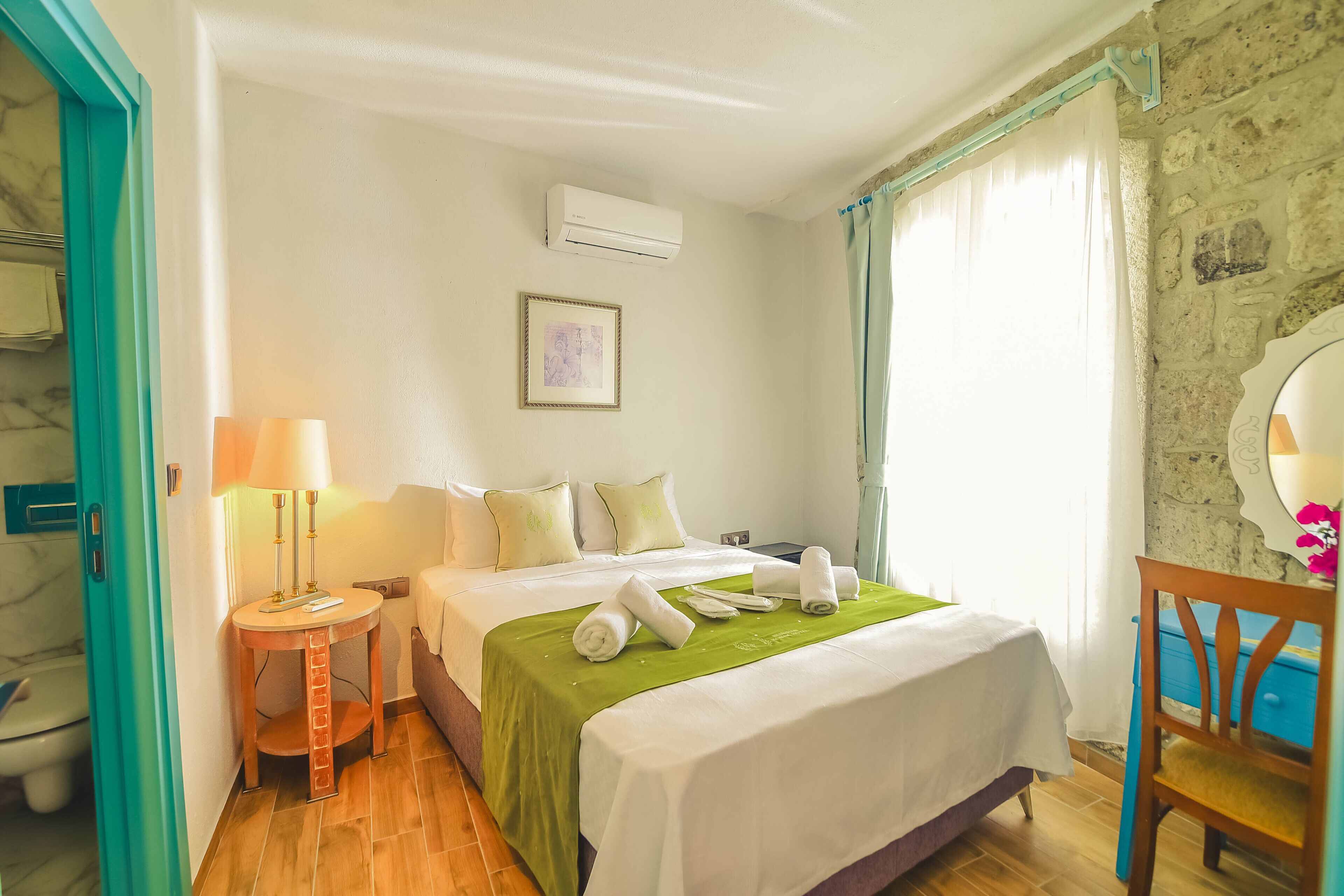 Habitación de hotel con cama, mesita de noche con lámpara, espejo y ventana con cortinas.
