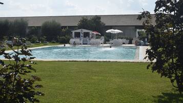 Una piscina con sillas de descanso y sombrillas, rodeada de césped y un edificio con tejado de baldosas.