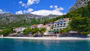 Ett hotell vid kusten med flera våningar, en strand i förgrunden och berg i bakgrunden.