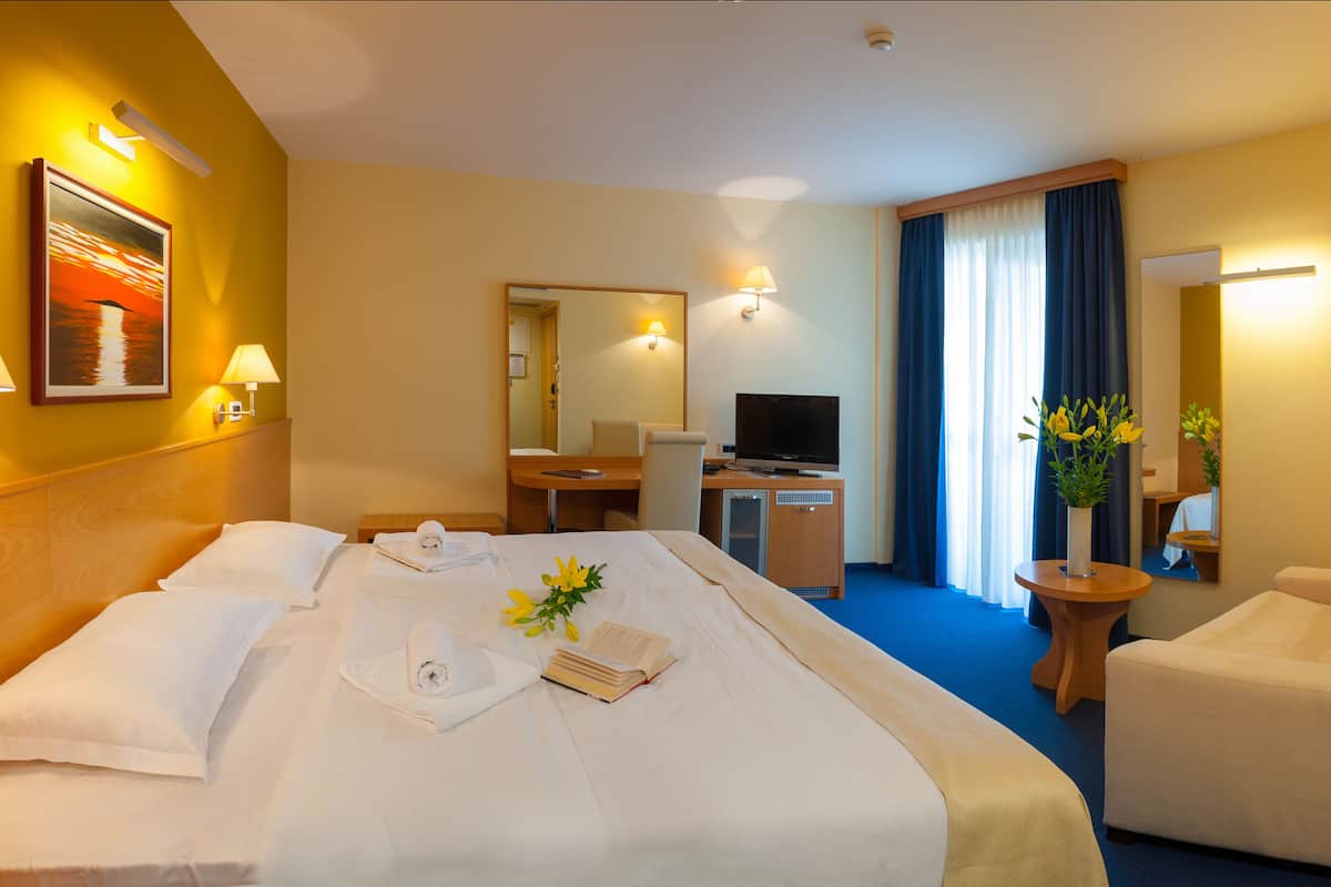 Quarto de hotel com uma cama grande, uma escrivaninha com computador, uma cadeira e uma mesinha de centro com um vaso de flores.
