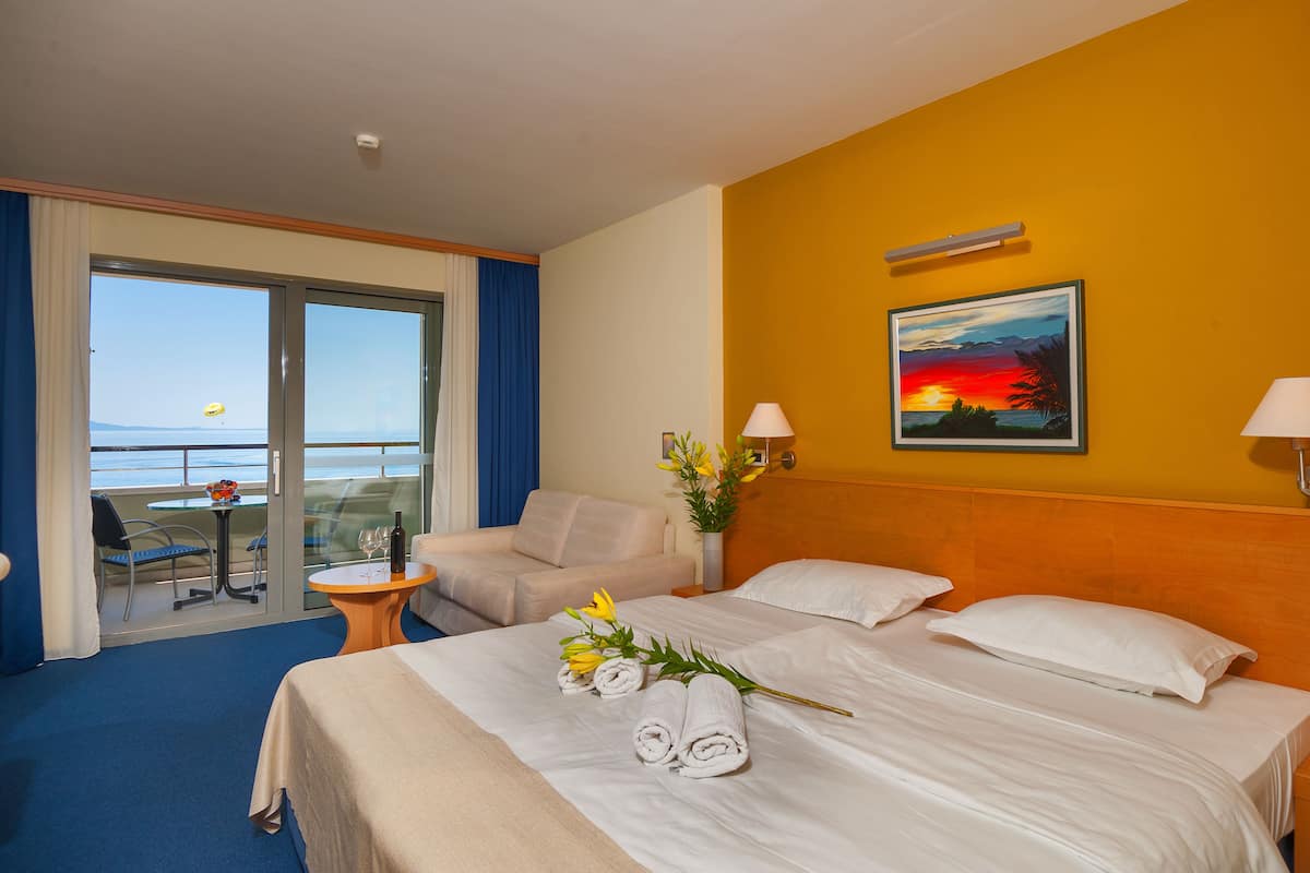 Quarto de hotel com cama, duas almofadas, mesa de cabeceira, vaso com flores, quadro na parede, janela com vista para o mar e varanda com mesa e cadeiras.