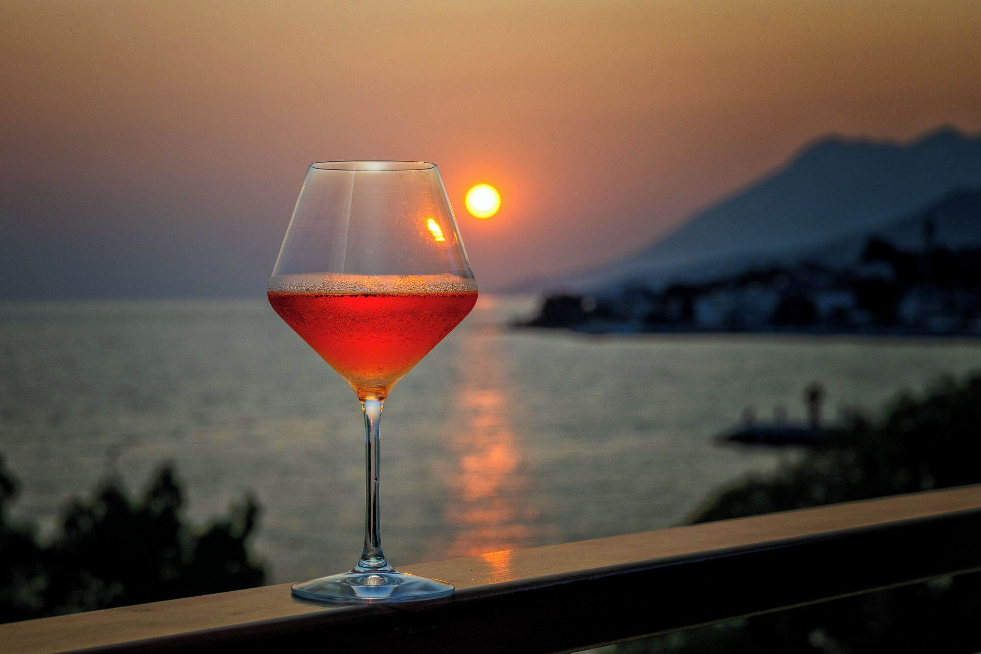 A glass of red wine on a balcony with a sunset and mountains in the background.