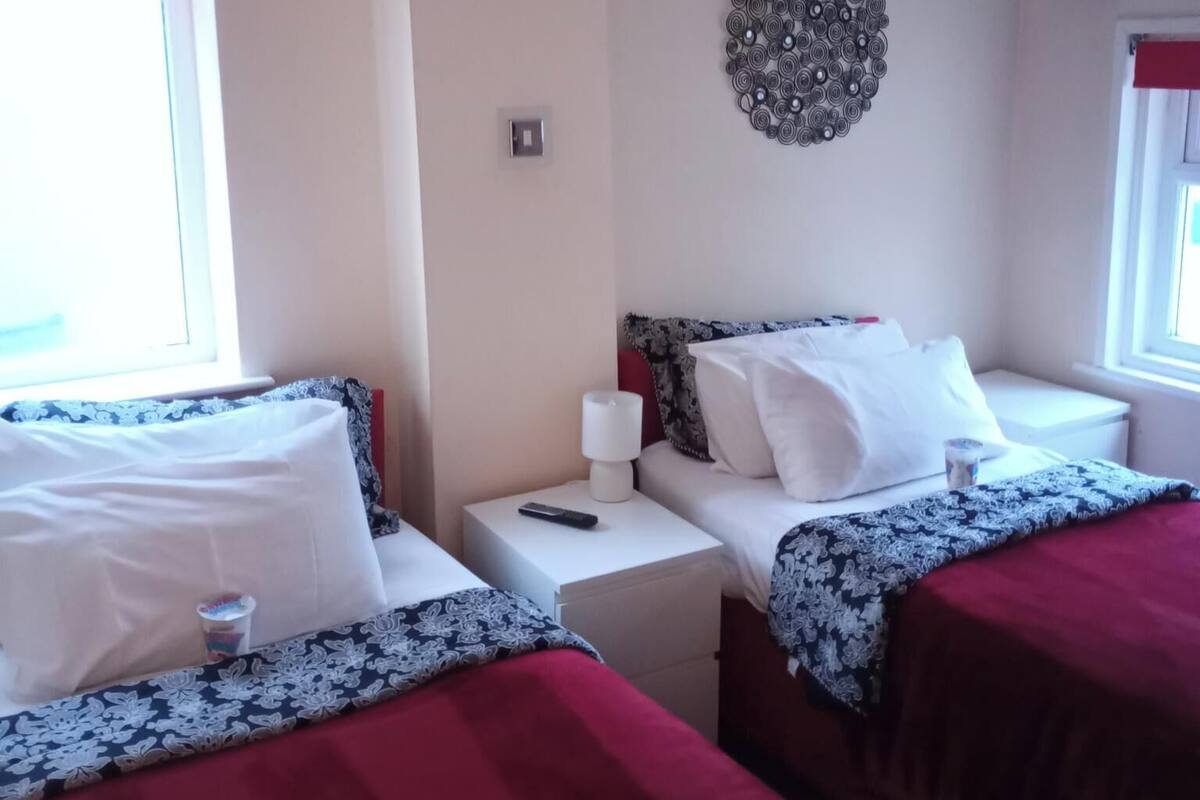 A hotel room with two beds, a bedside table with a lamp, a wall decoration, and a window with a red curtain.