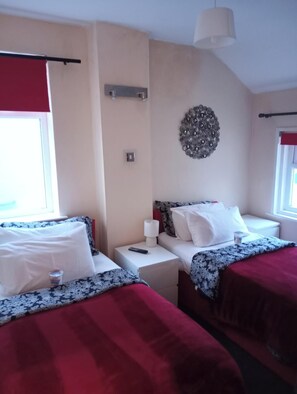 A hotel room with two beds, a bedside table with a lamp, a wall decoration, and a window with a red curtain. - Aparthotel Blackpool (Blackpool)
