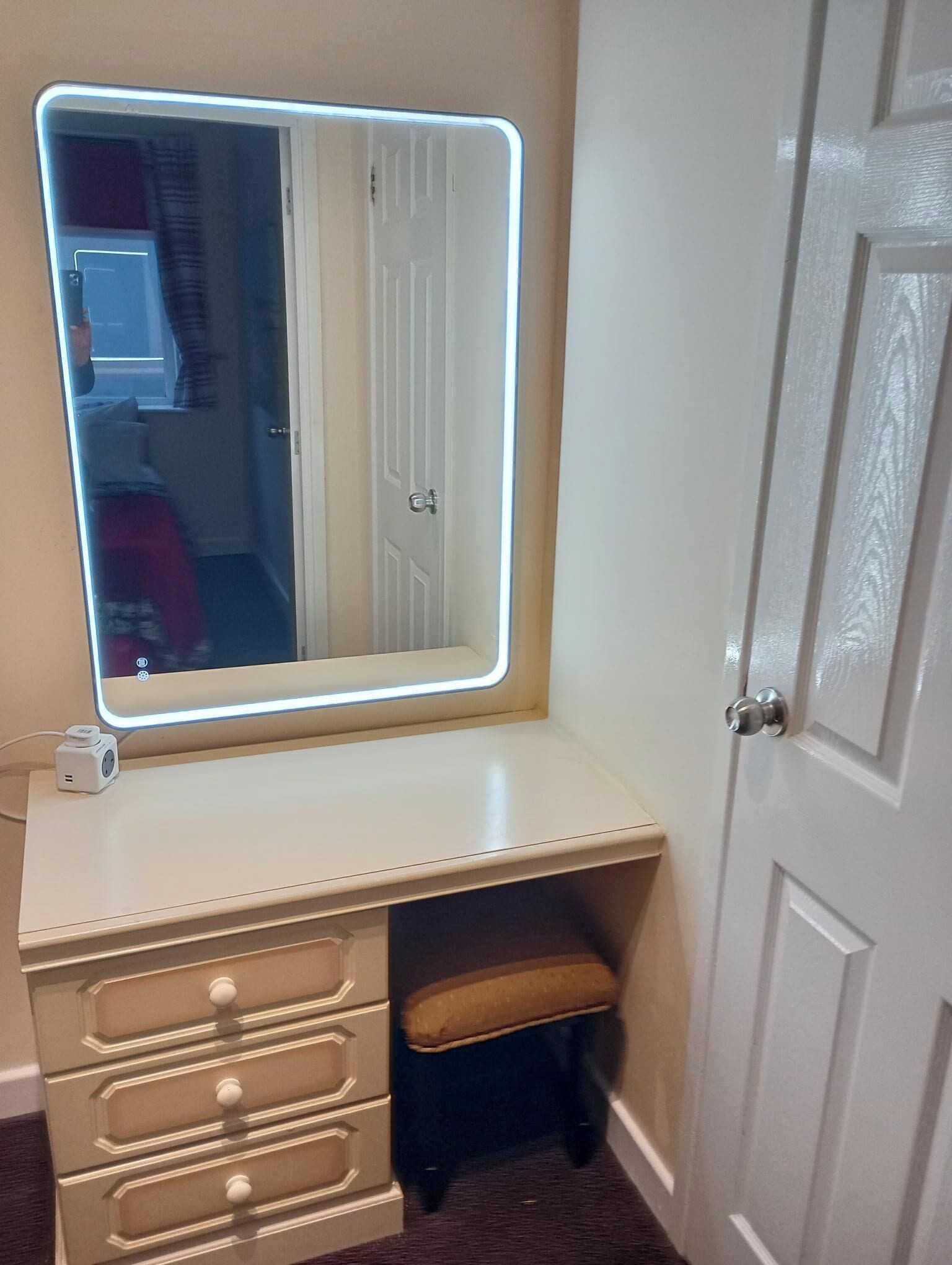 Un tocador con un espejo grande y un marco iluminado, un taburete pequeño y un mueble con pomos.