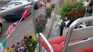 Un balcon doté de balustrades en métal, de coussins rouges et de plantes en pot fleuries. Des drapeaux sont fixés aux balustrades.