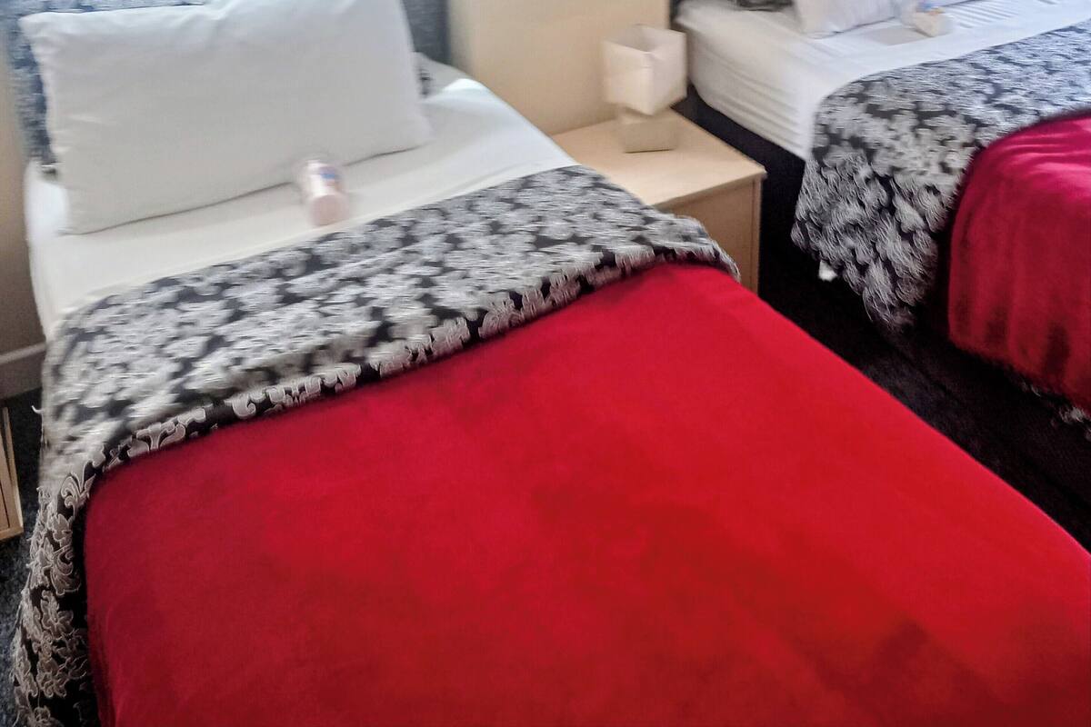 A hotel room with two beds, each with a patterned bedspread and white pillows. There is a bedside table with a tissue box and a lamp. A painting of flowers hangs on the wall.