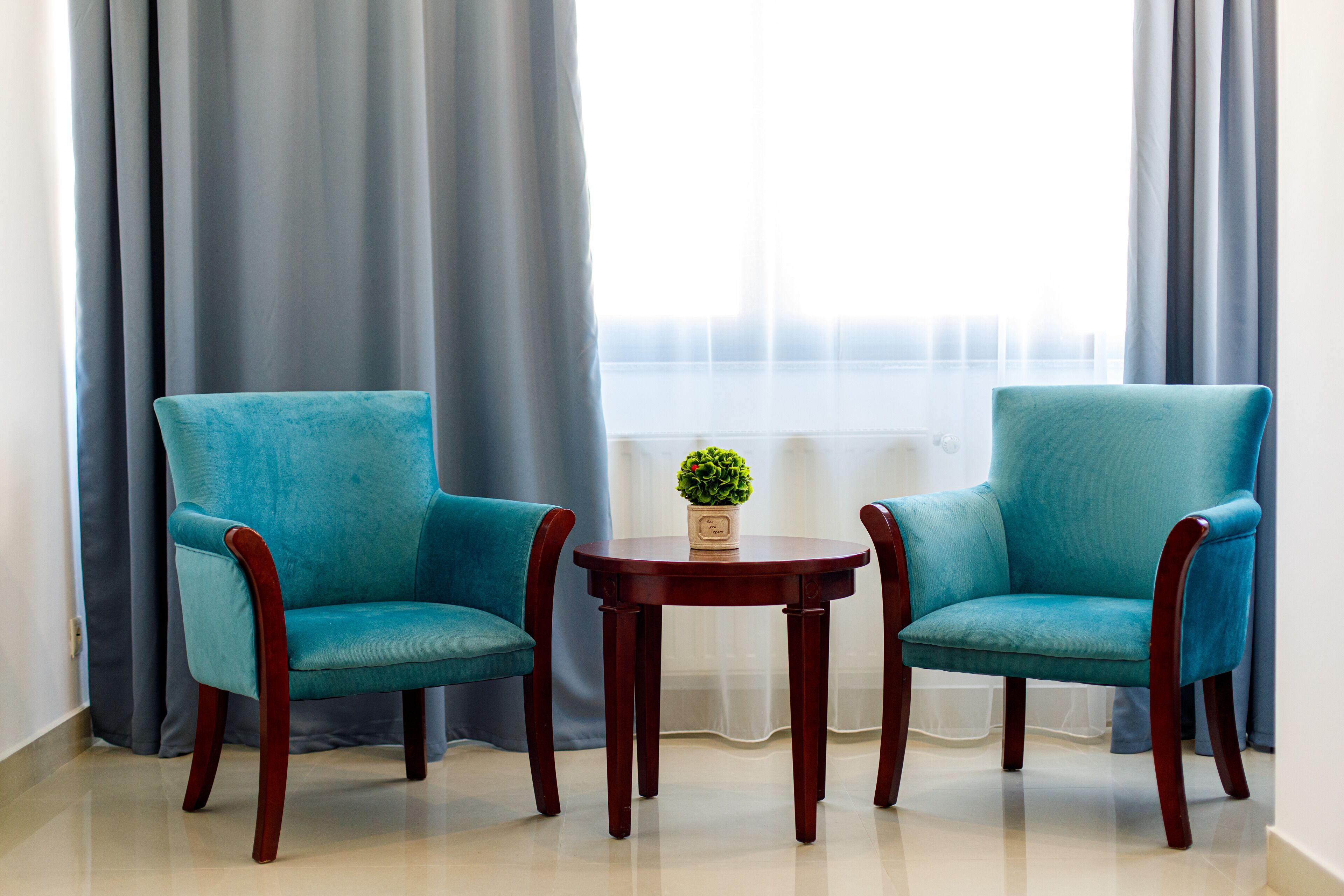 Two teal armchairs and a small wooden table with a potted plant in a room with large windows and grey curtains.