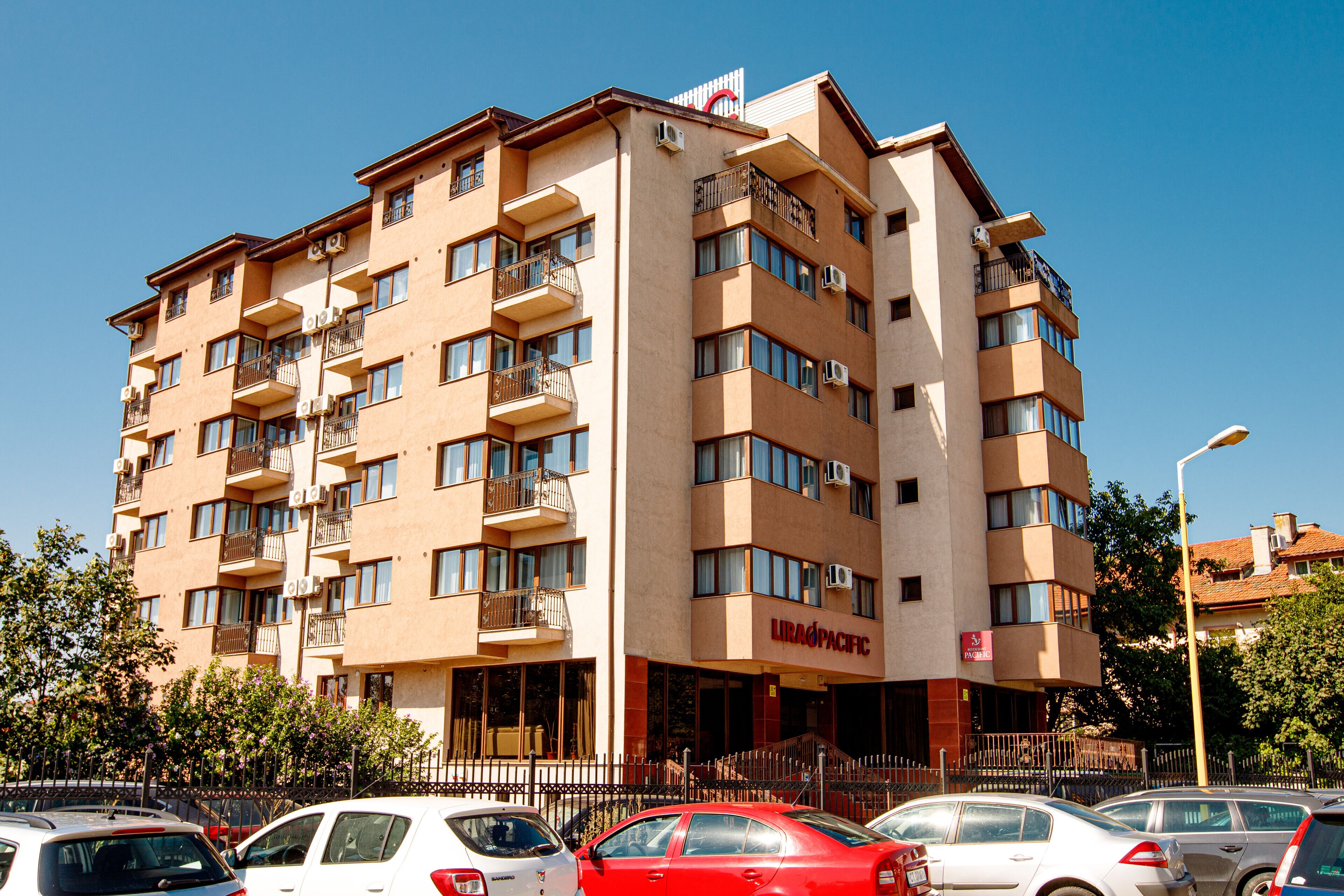 Un hotel de varios pisos con balcones, un letrero que dice "LIRAG PACIFIC" y varios coches estacionados al frente.