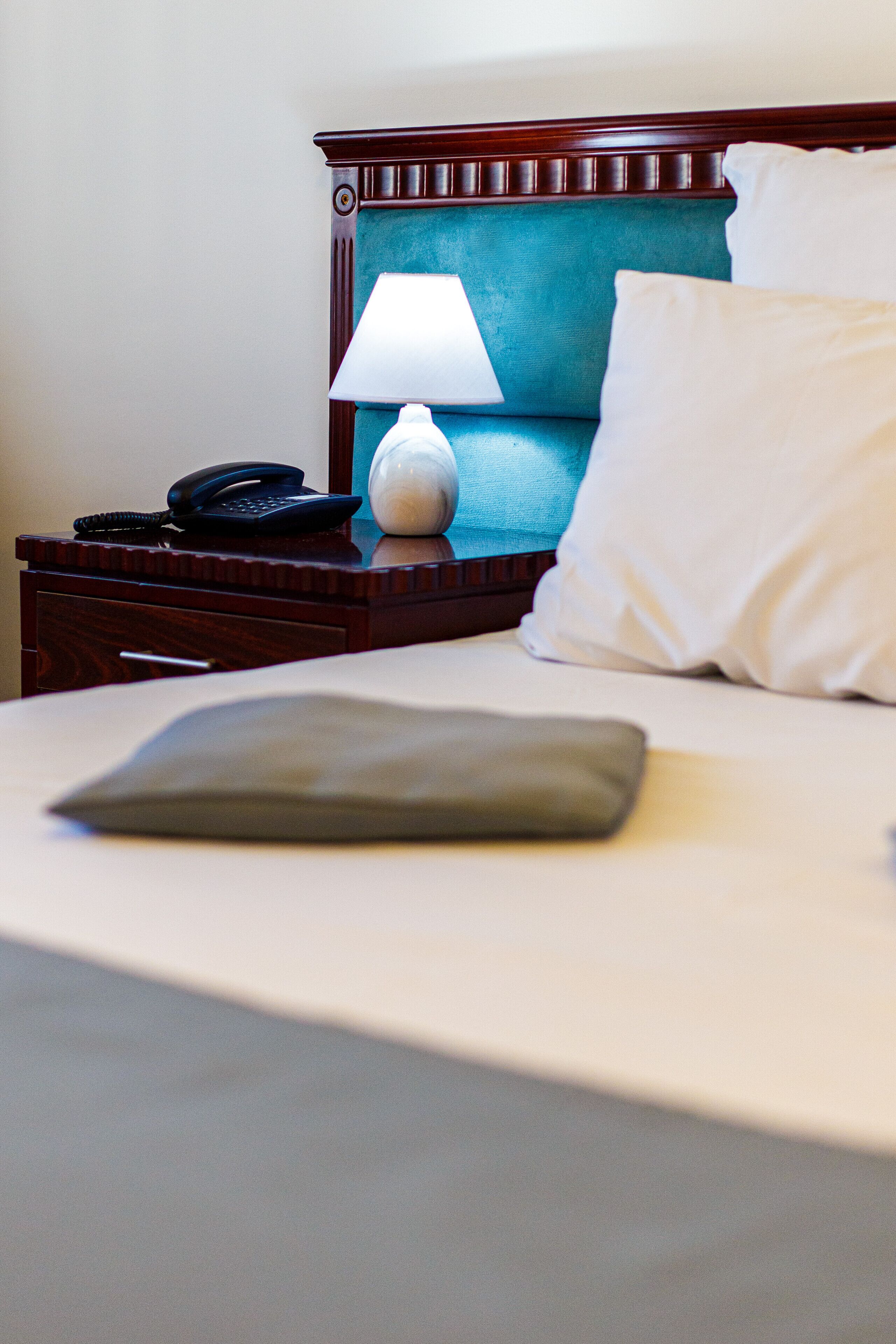 Een net opgemaakt bed met een kussen, een bedlampje en een telefoon op een houten nachtkastje.
