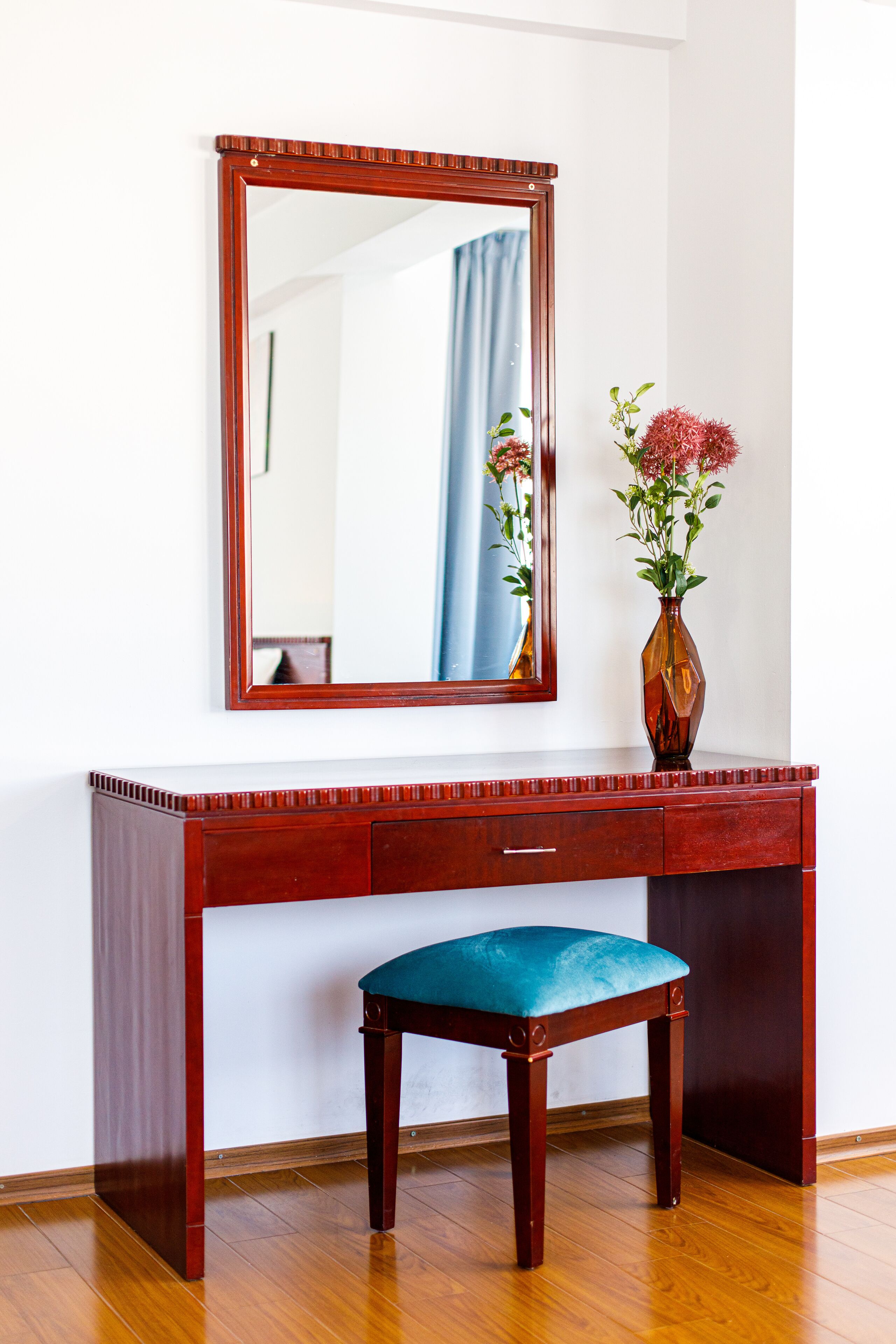 Een houten bureau met een spiegel, een vaas met bloemen en een blauwe fauteuil.
