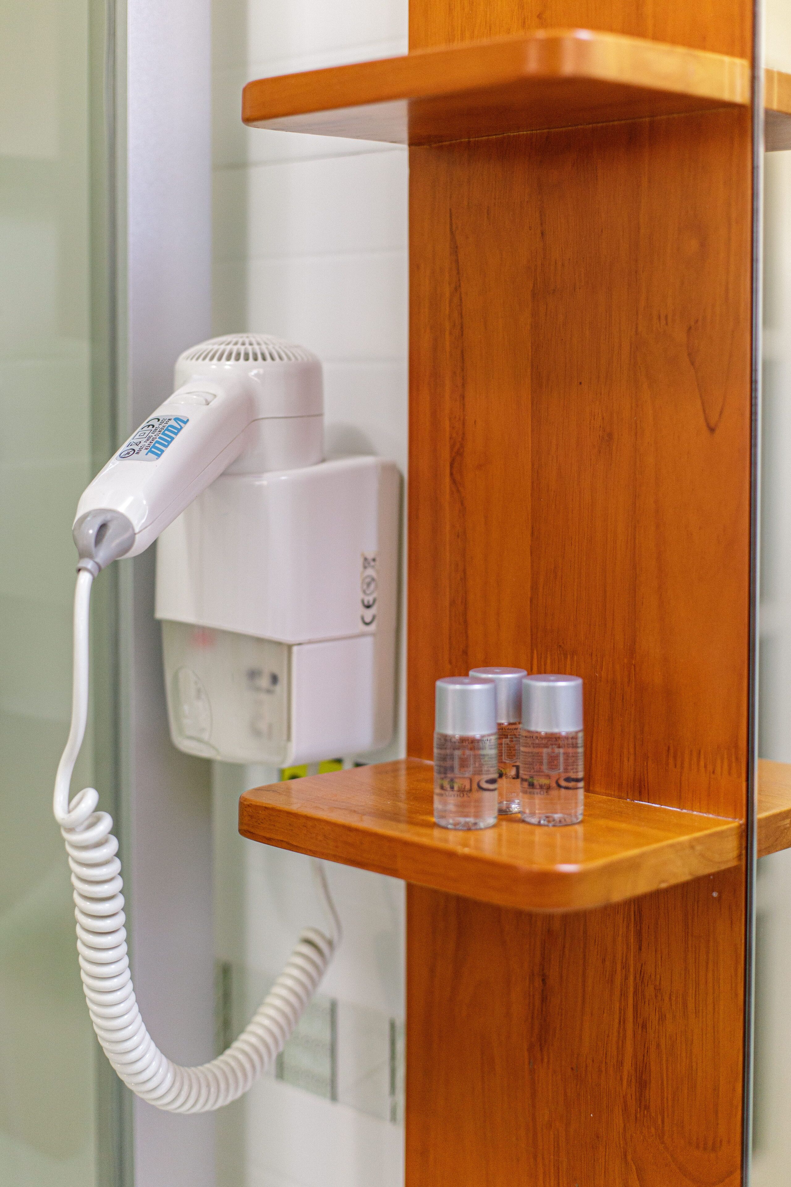 A hairdryer on a white wall, a wooden shelf with three bottles, and a white electrical outlet.