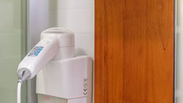 A hairdryer on a white wall, a wooden shelf with three bottles, and a white electrical outlet.