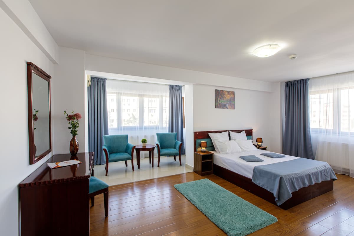 Habitación de hotel con una cama grande, dos sillones, una mesita con un florero y un escritorio con un espejo y un florero.