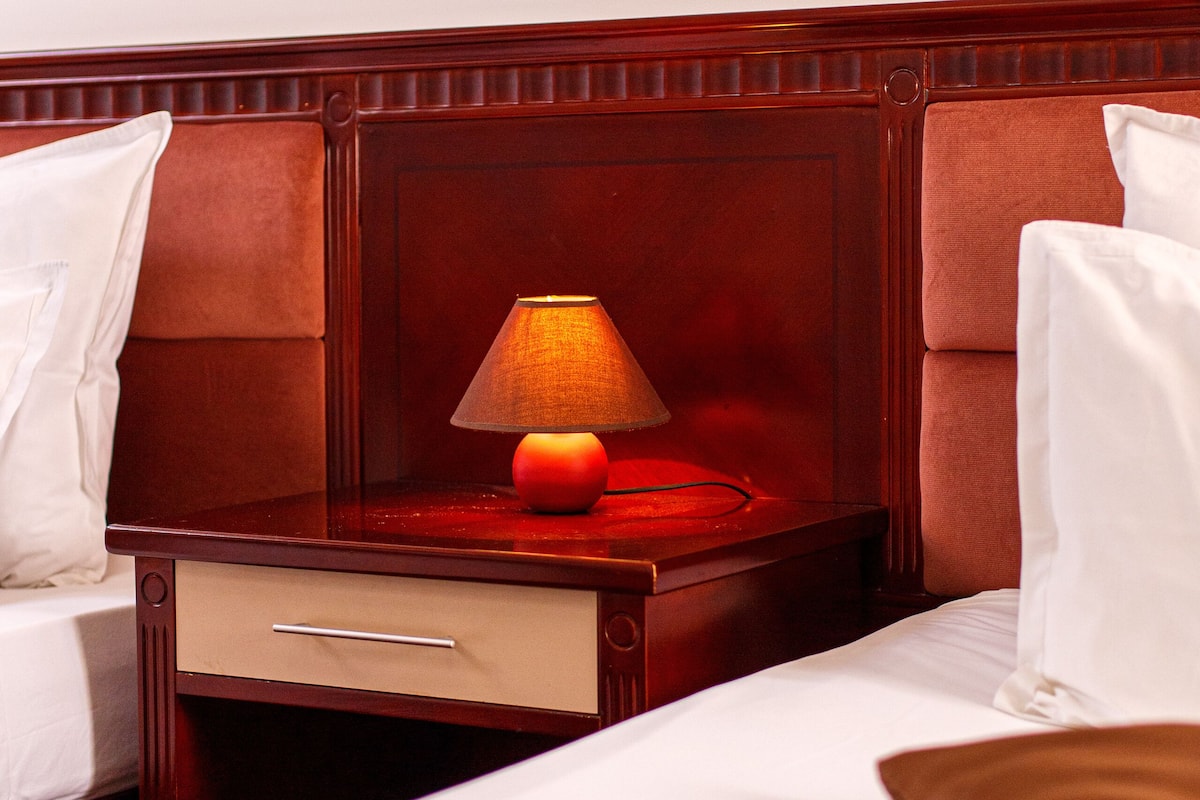 Habitación de hotel con cabecera de madera, mesita de noche con lámpara y cama con ropa de cama blanca.