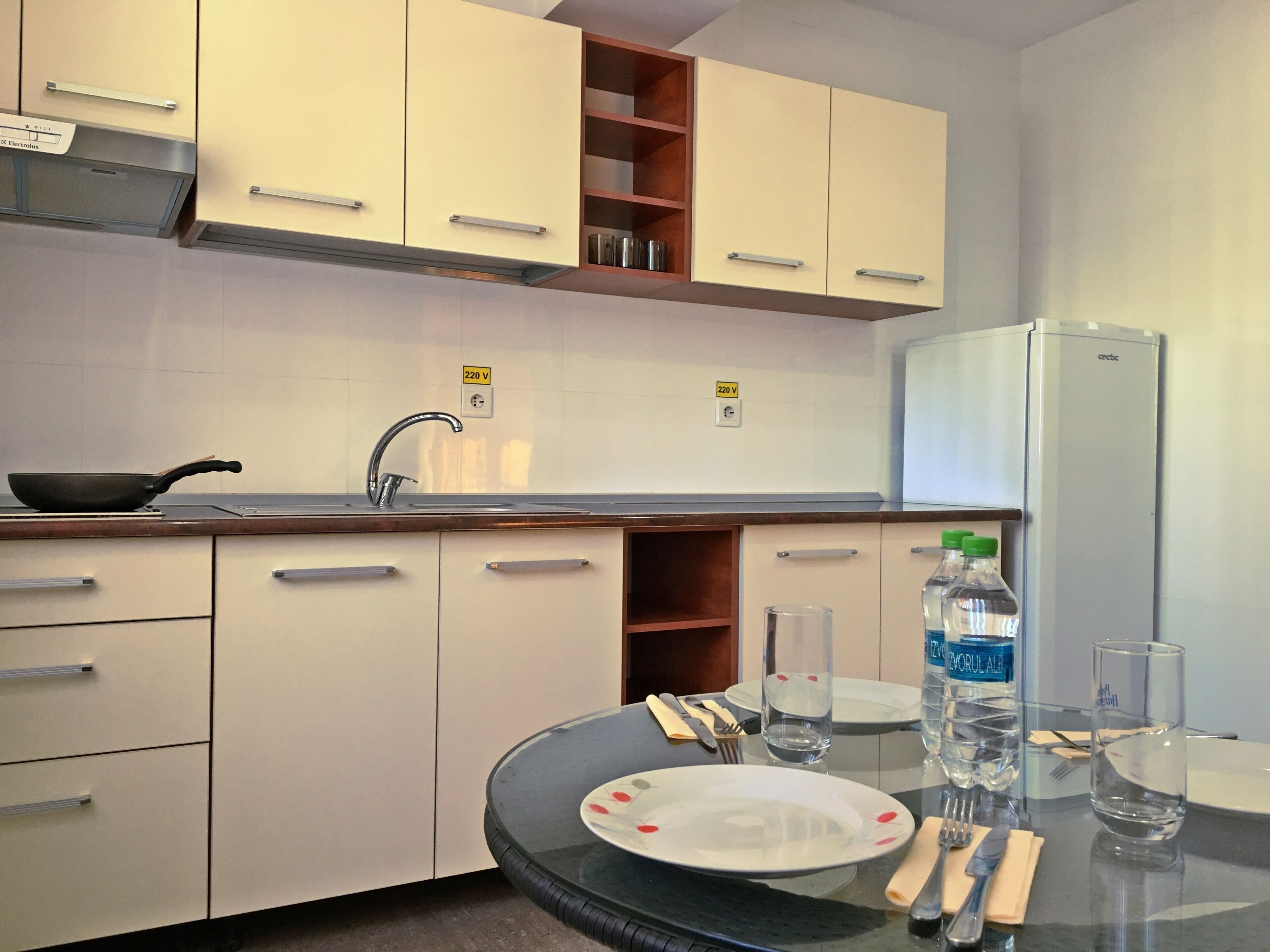 Eine moderne Küche mit weißen Schränken, einer Edelstahl-Dunstabzugshaube, einem Kühlschrank und einem Esstisch, der mit Tellern, Gläsern und Besteck gedeckt ist.