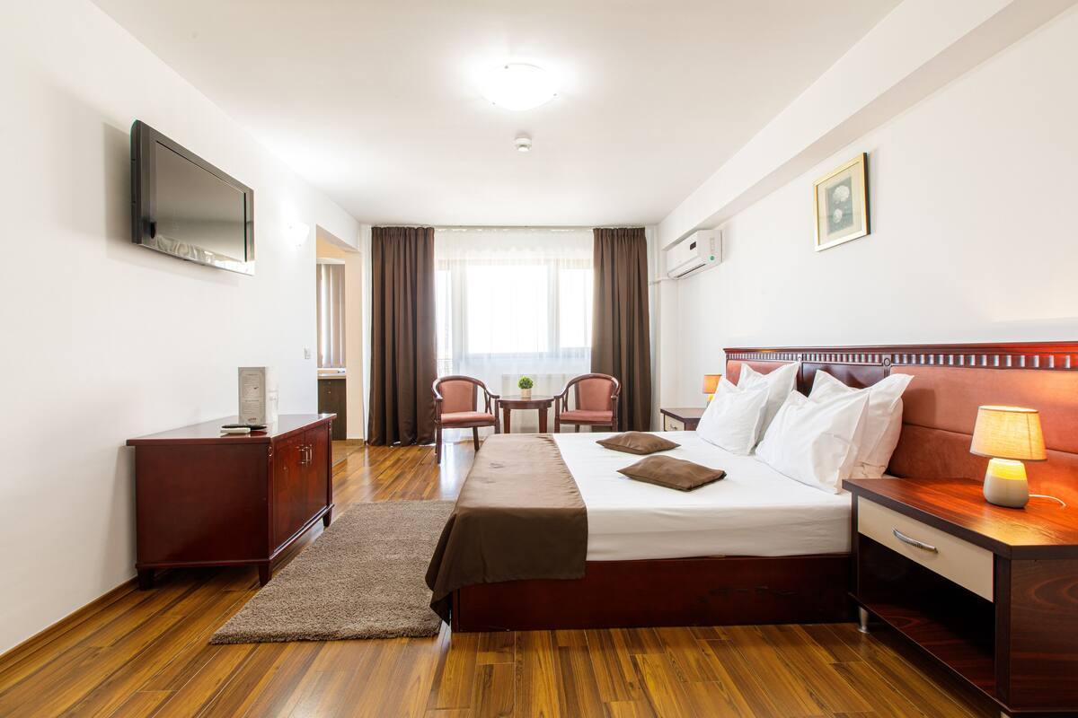 Habitación de hotel con cama, mesita de noche, televisión, escritorio, sillas y ventana con cortinas.