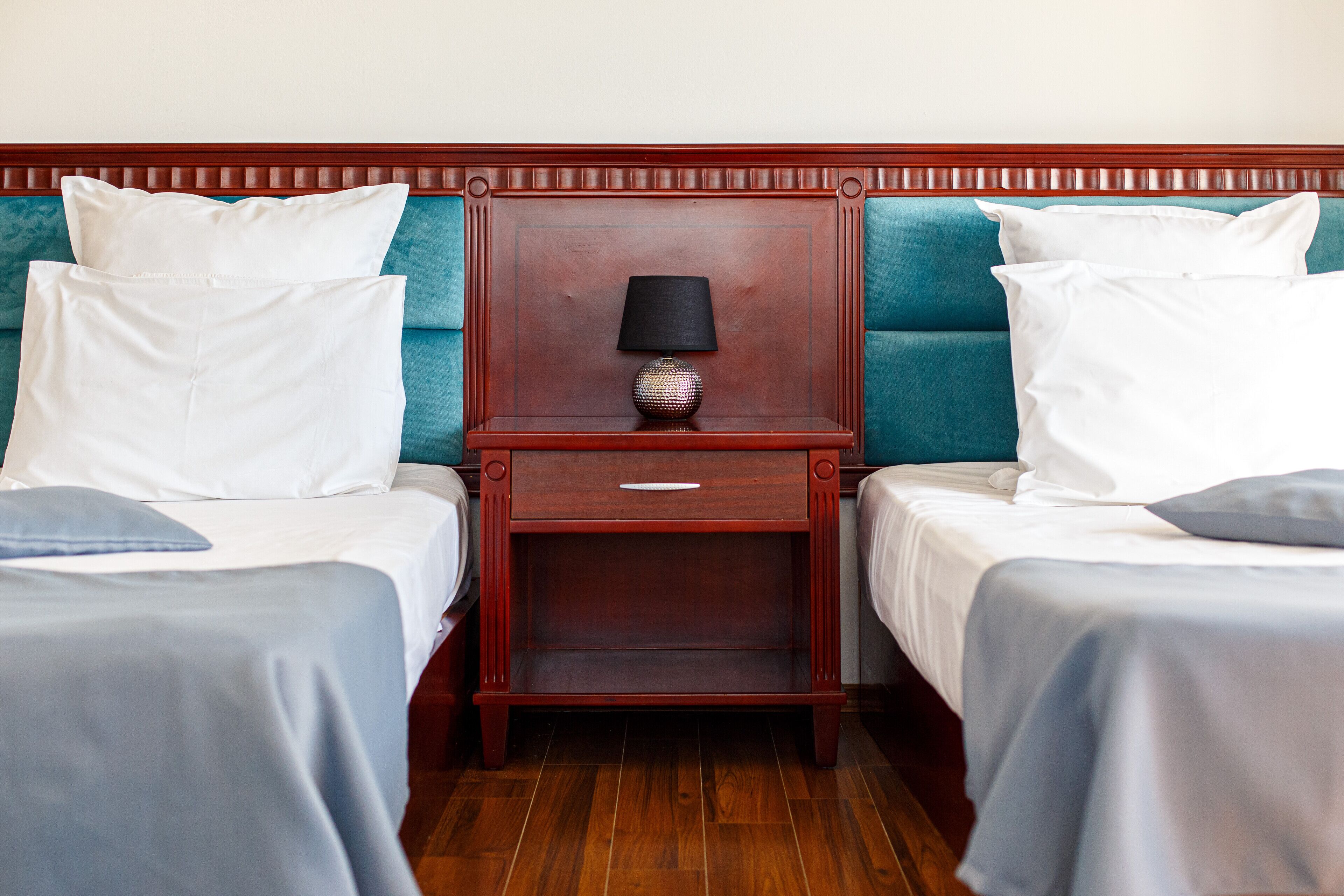 Habitación de hotel con dos camas, una mesita de noche con lámpara y cabeceros tapizados en color verde azulado.