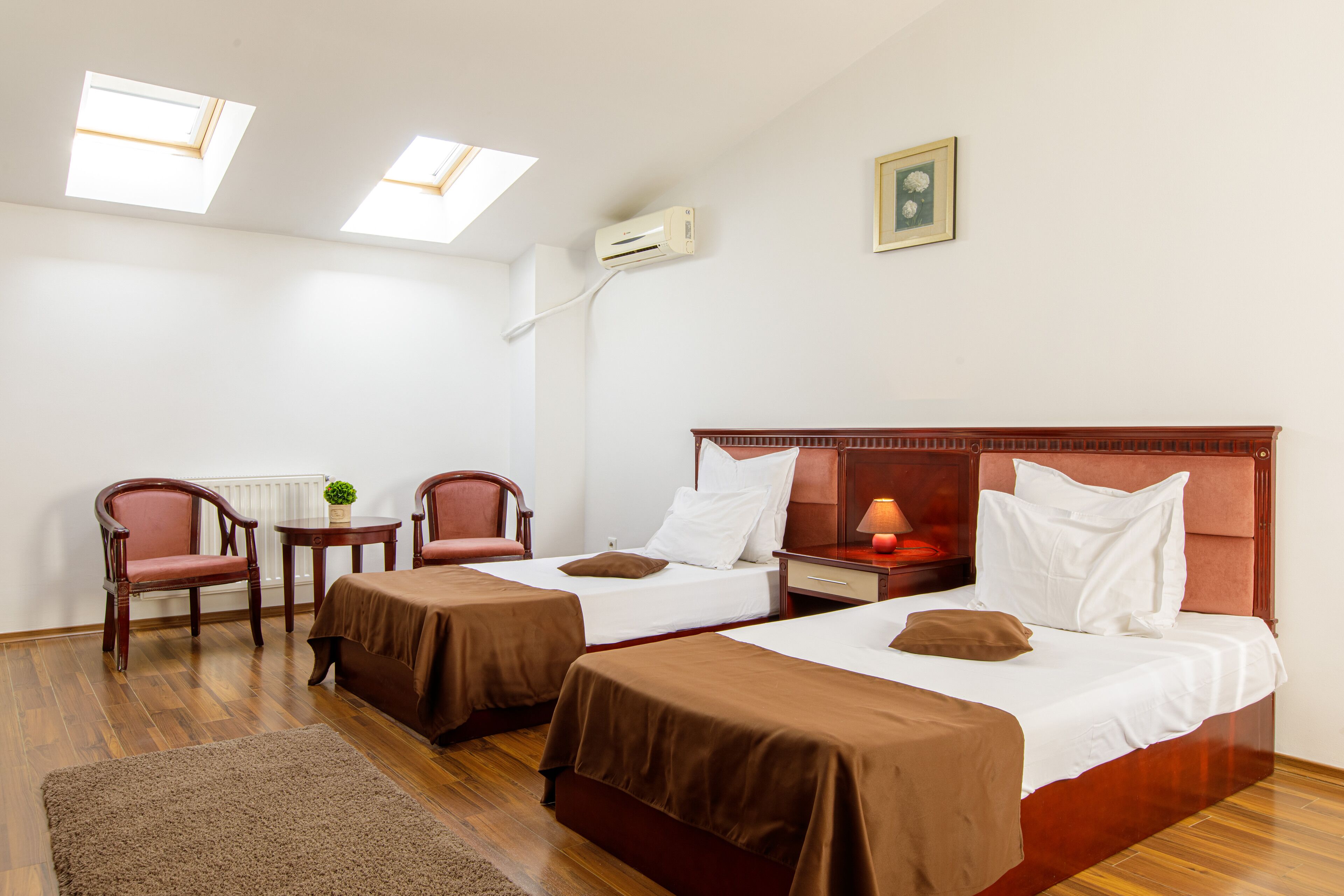 Habitación de hotel con dos camas, piso de madera, una mesita con sillas y una ventana con tragaluces.