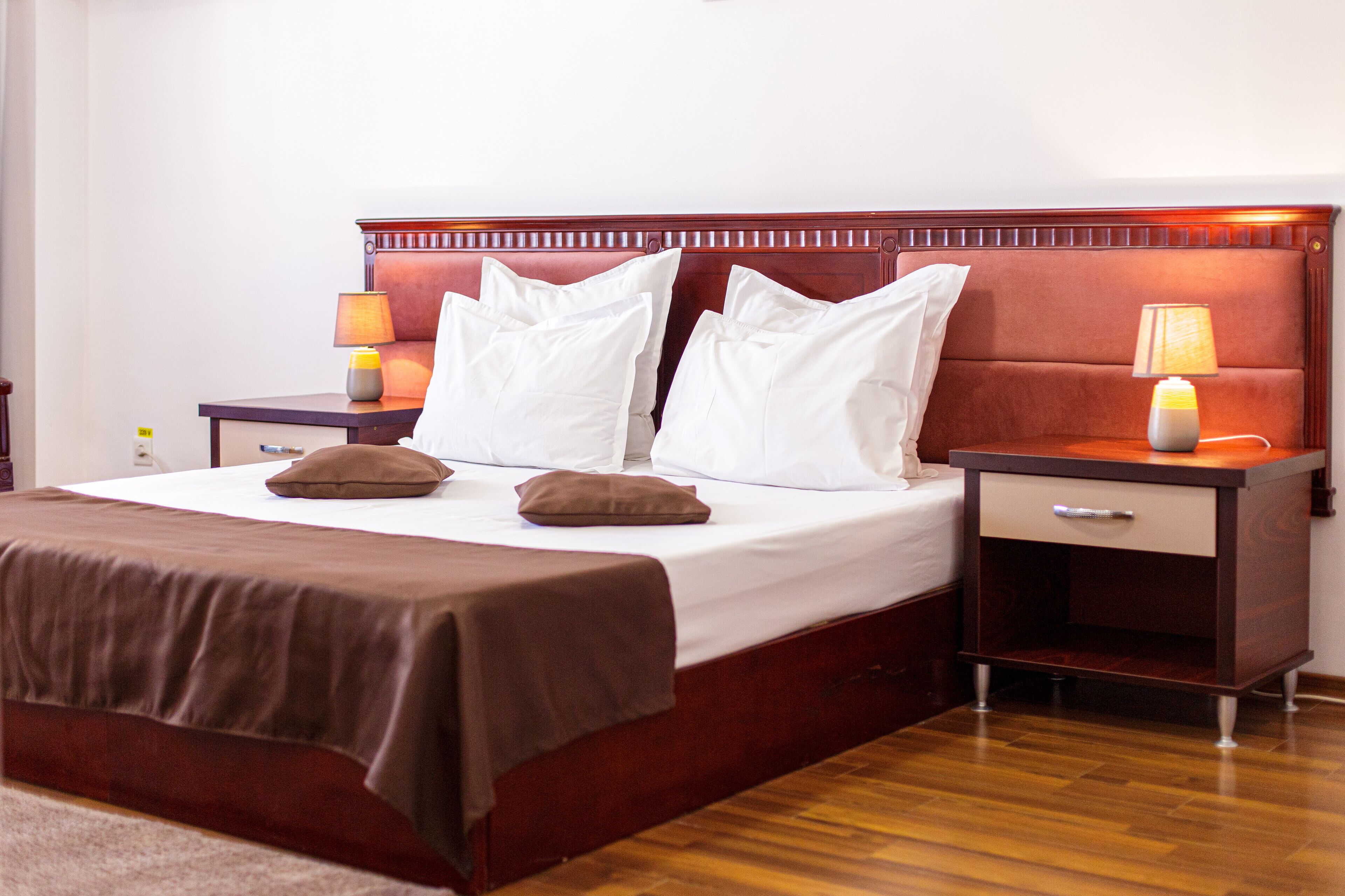 Una cama bien tendida con un cabecero marrón, dos mesitas de noche con lámparas y suelo de madera.