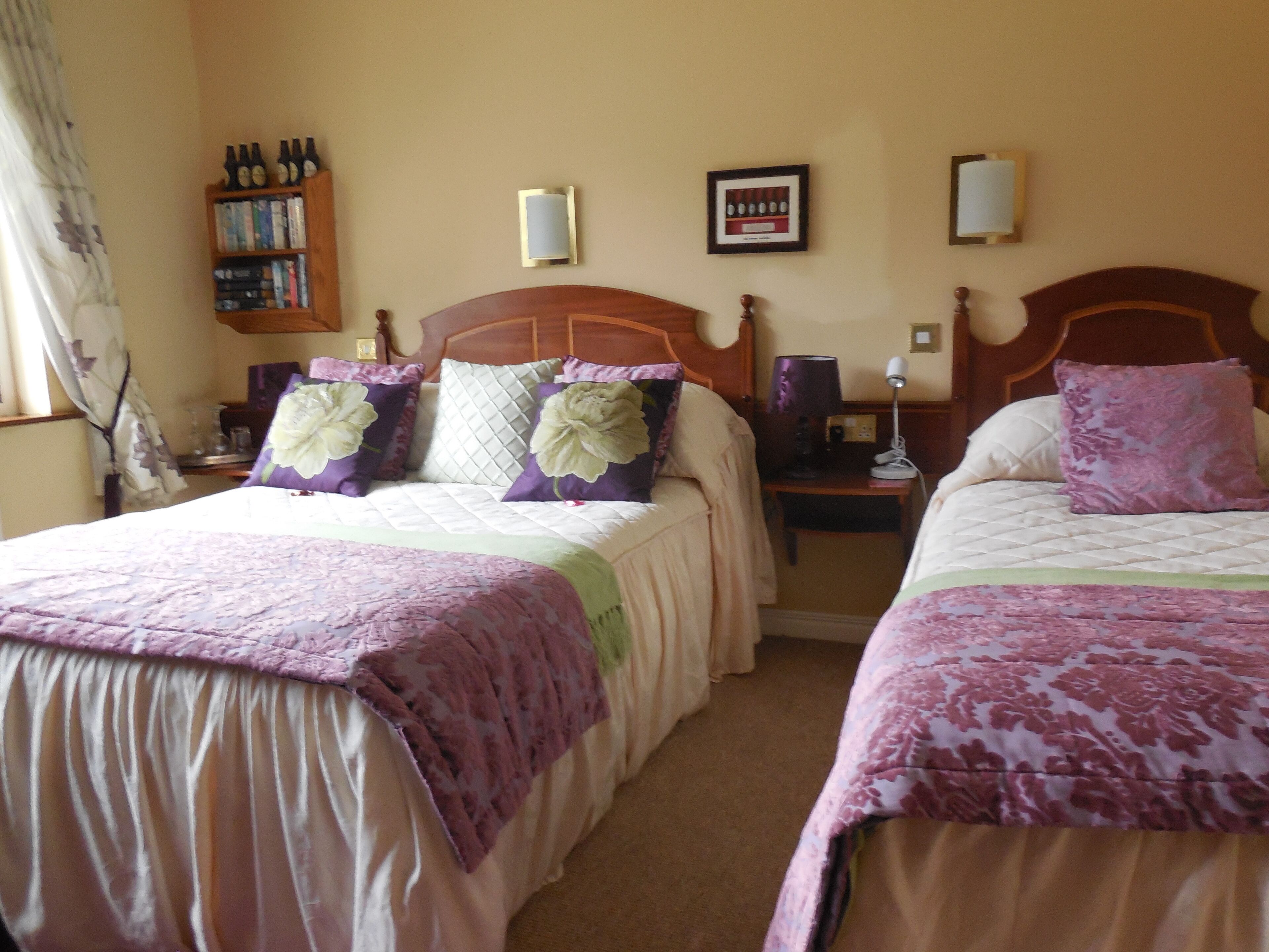 Ett hotellrum med två sängar, var och en med ett blommigt sängöverkast och matchande kuddar. Det finns en bokhylla med böcker och dekorativa föremål, en inramad tavla på väggen och ett nattduksbord med en lampa.