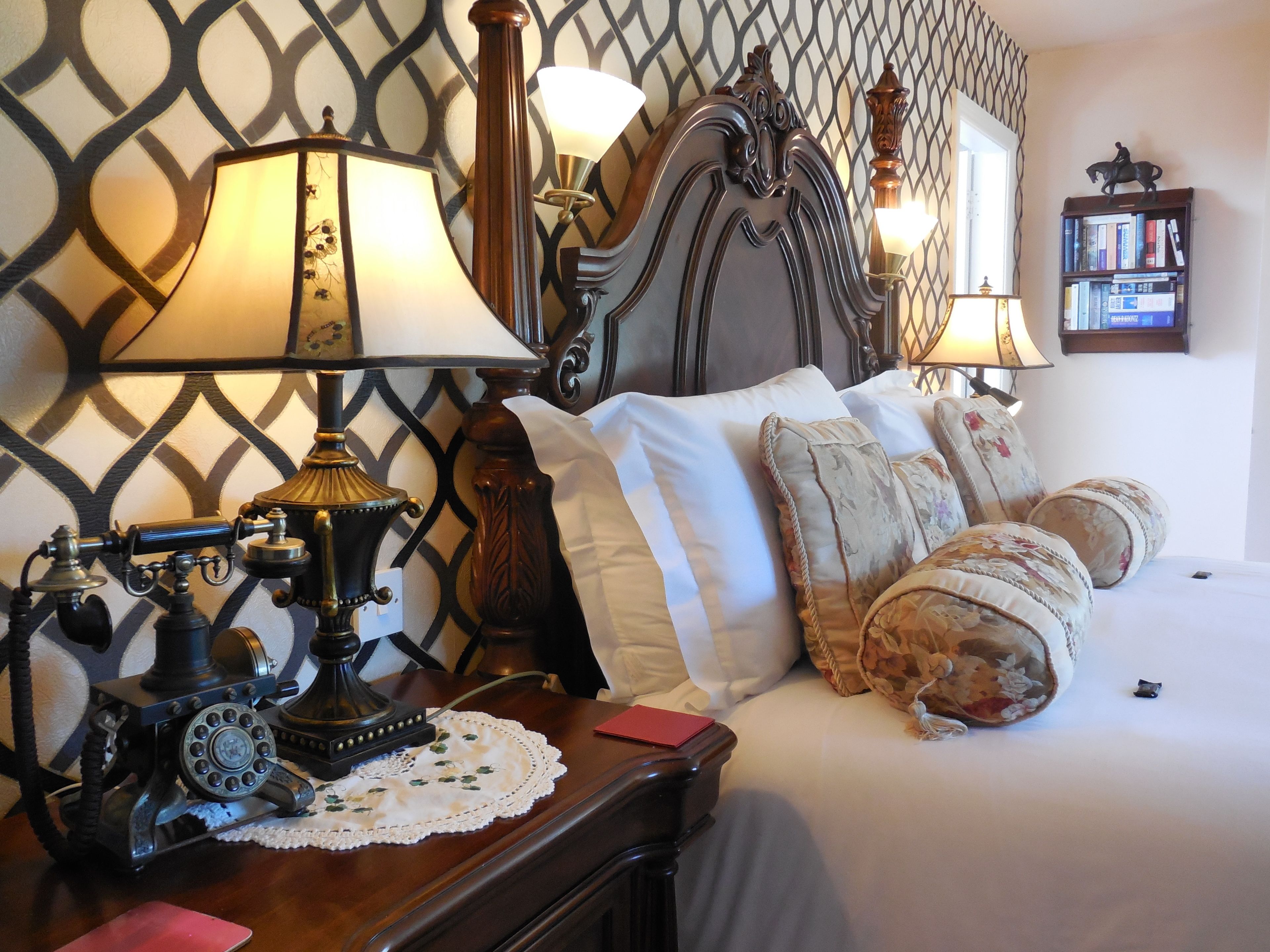 A neatly made bed with floral pillows, a vintage telephone on a wooden nightstand, and a wall with a geometric pattern.
