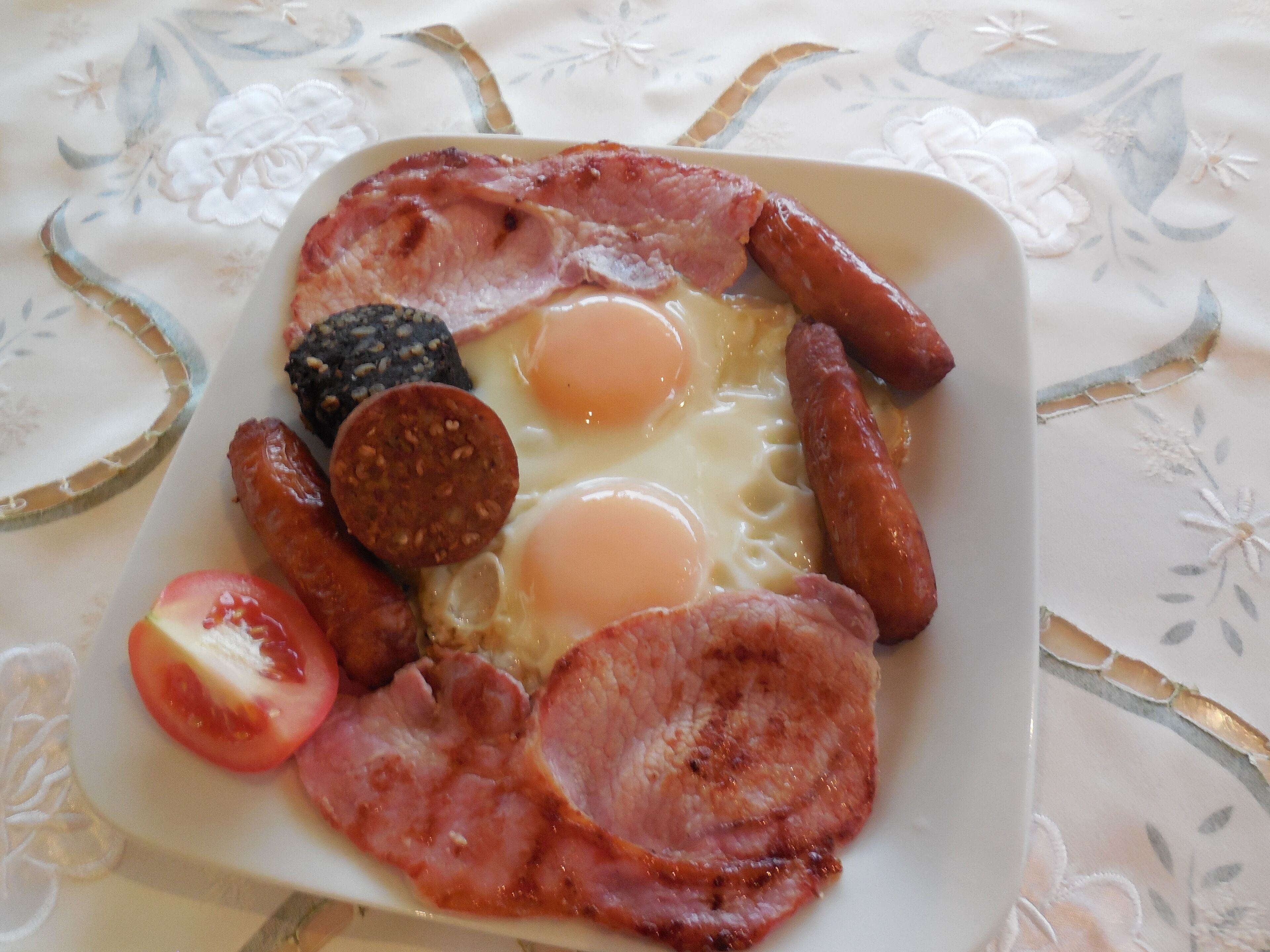 A breakfast plate with eggs, sausages, bacon, and a slice of tomato.