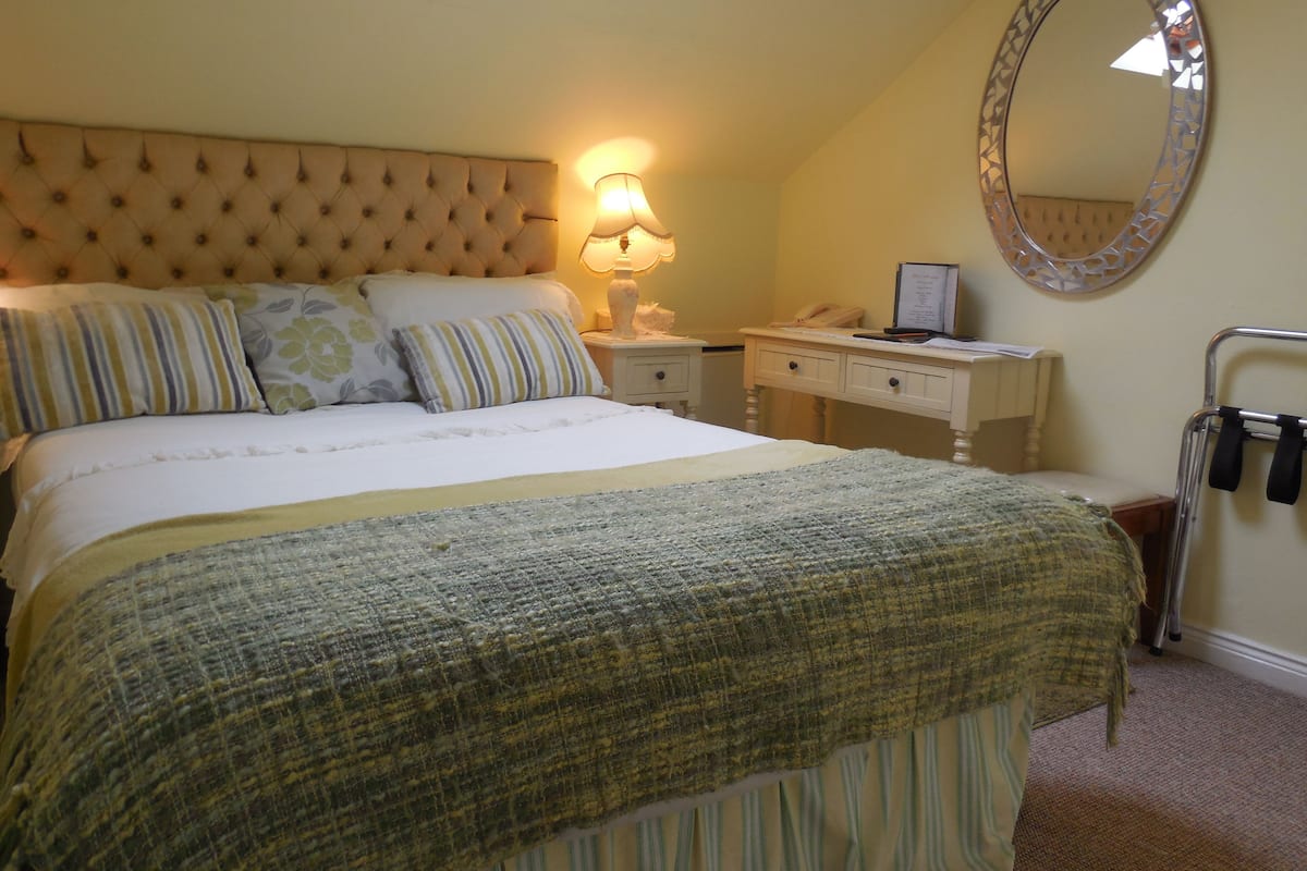 A bedroom with a tufted headboard, a bed with striped bedding, a bedside table with a lamp, a desk with a mirror, and a wheelchair.