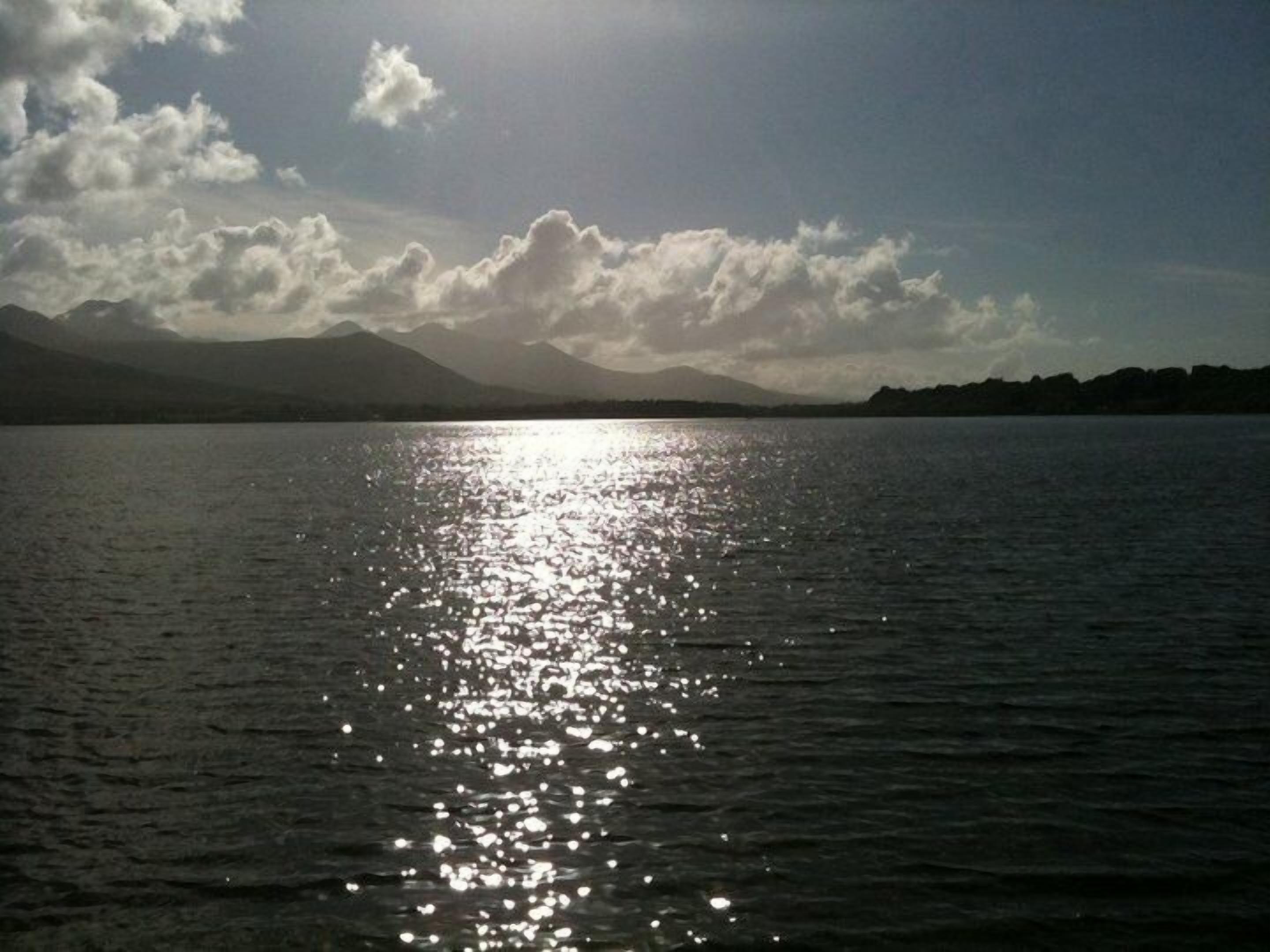 A calm lake with sunlight reflecting off the water's surface, surrounded by mountains and a partly cloudy sky.