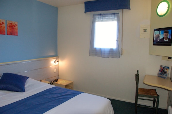 Habitación de hotel con cama, mesita de noche con lámpara, ventana con cortina, un taburete pequeño y televisor montado en la pared.
