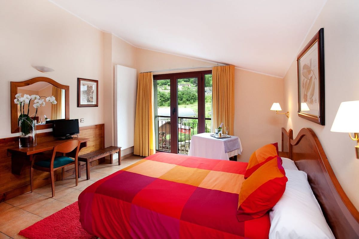 A hotel room with a bed, a desk with a computer, a chair, a small table with a lamp, and a window with curtains.