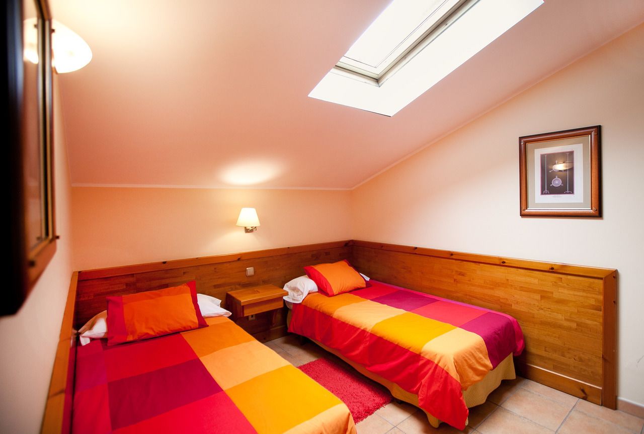 A small, cozy room with two single beds, a nightstand, a wall-mounted lamp, and a framed picture on the wall.